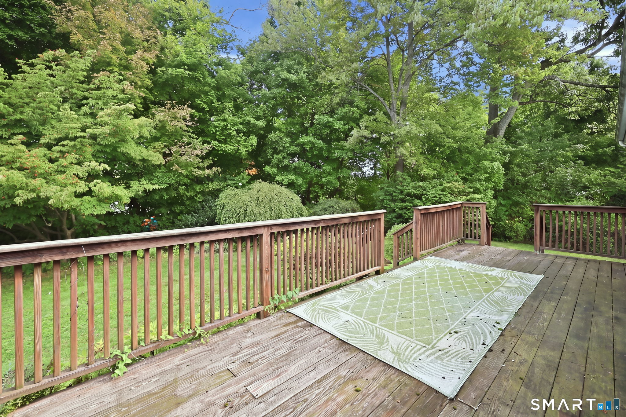 Wooden deck with a pale green patterned rug, overlooking a lush, tree-filled yard.
