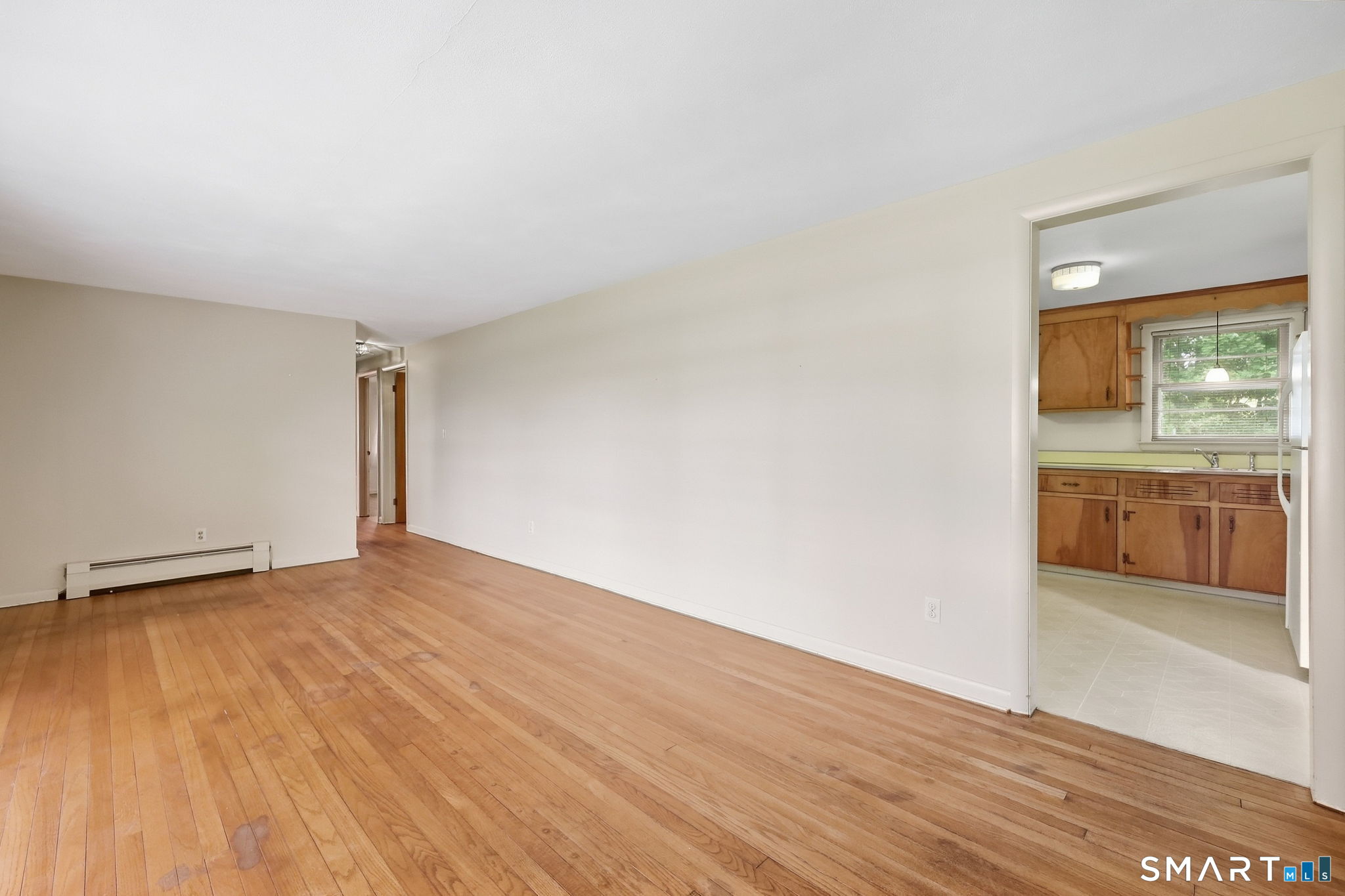Vacant living room with hardwood floors and a view into the wood-cabinet kitchen.