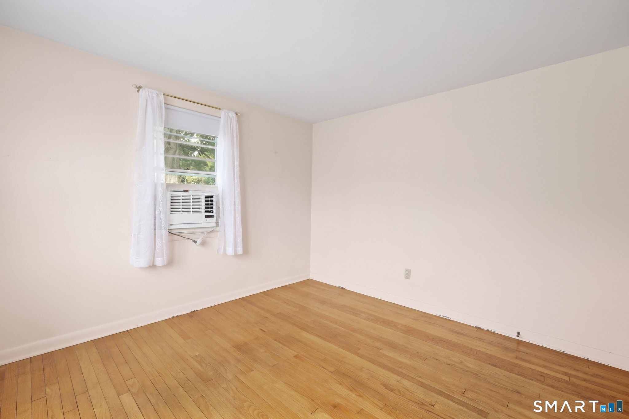 Empty room with hardwood floors and a window air conditioner behind white curtains.
