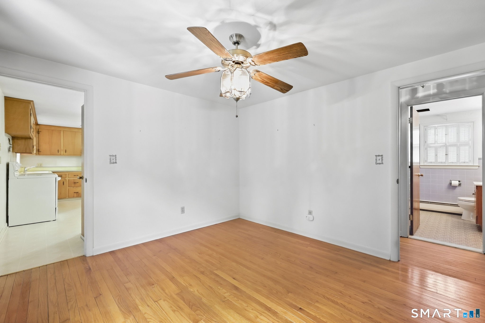 Empty room with hardwood floors and a ceiling fan, opening to the kitchen and bathroom.