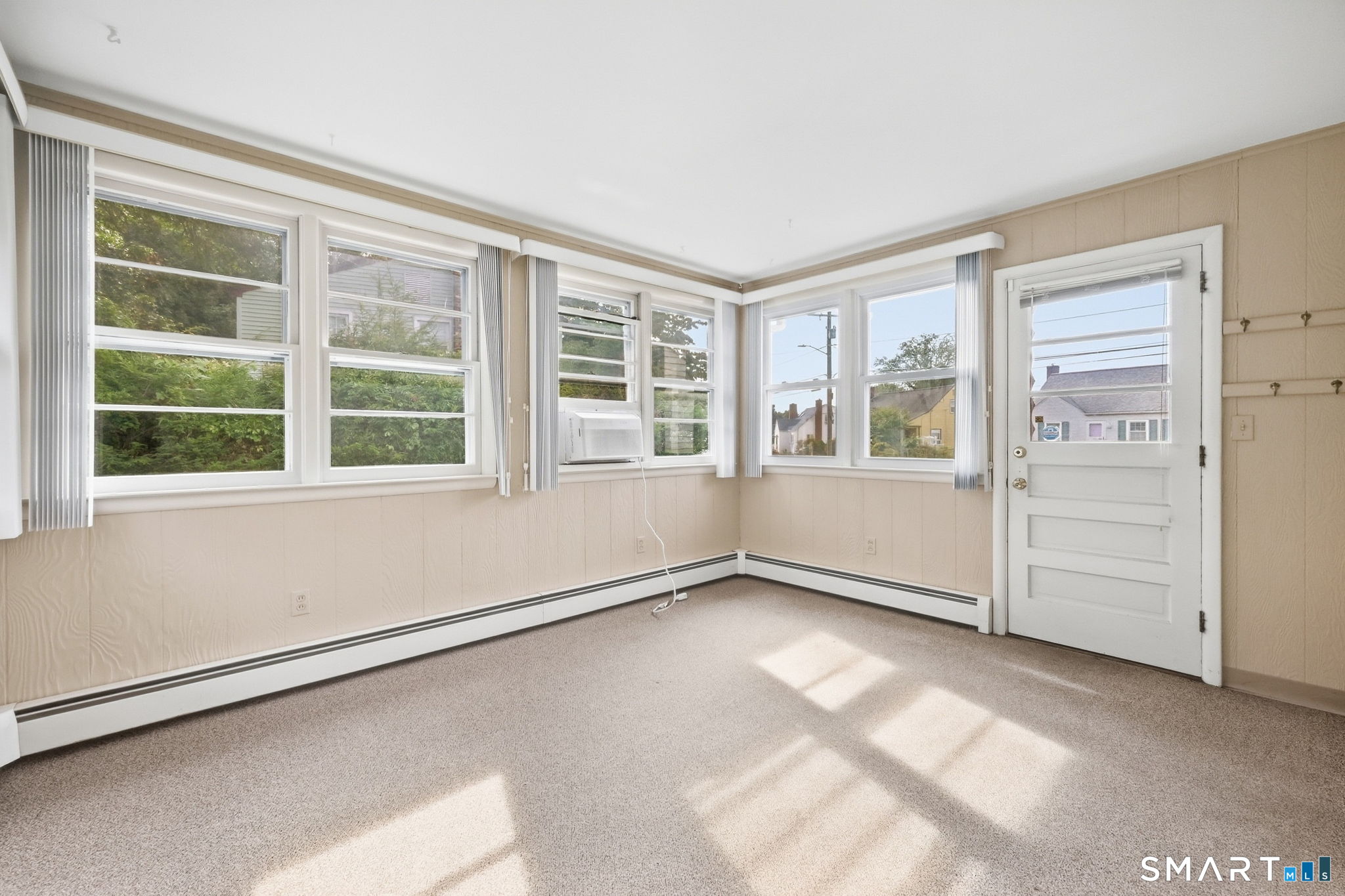 Empty carpeted room with wraparound windows, vertical blinds, and a white exterior door.