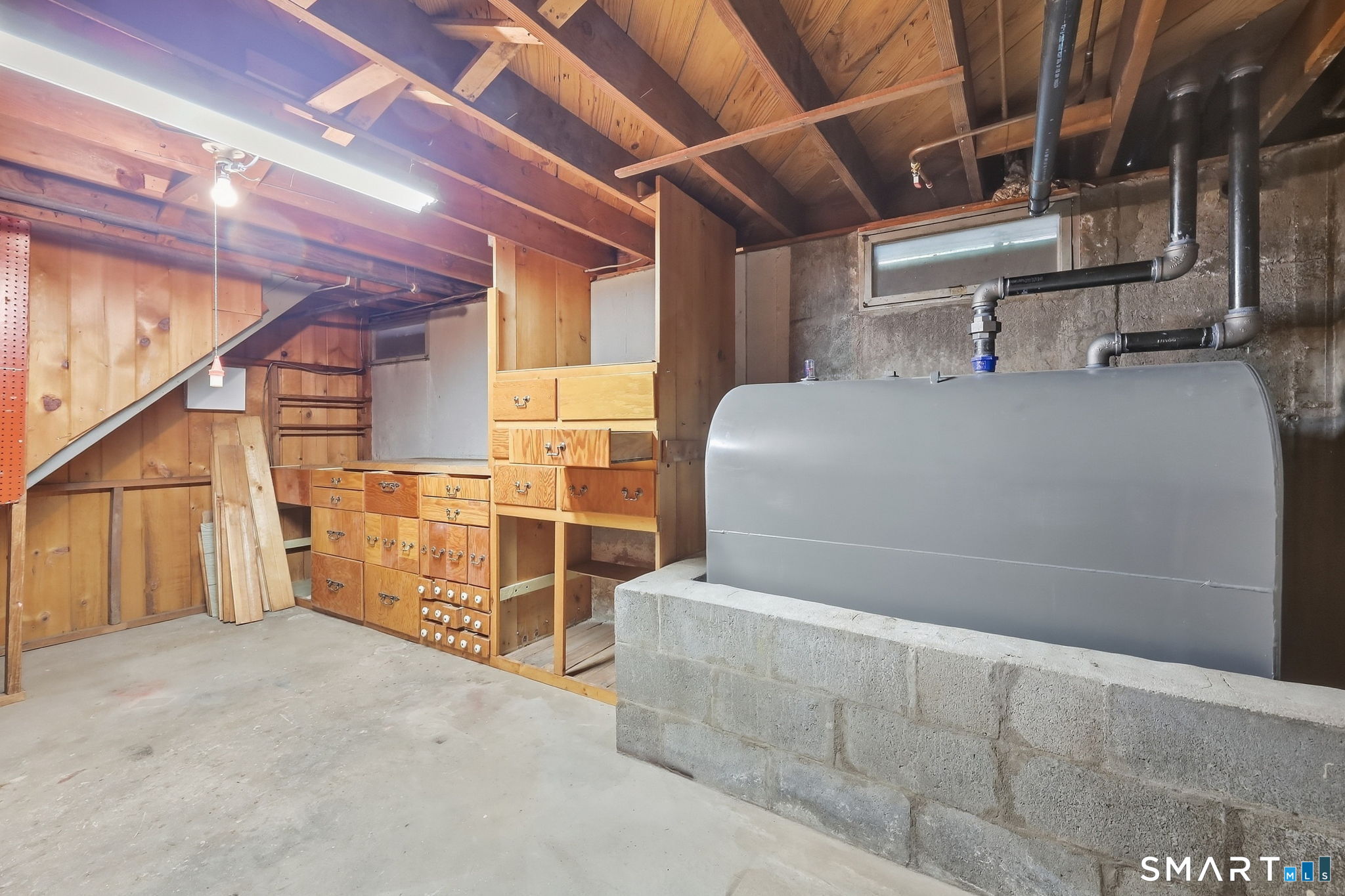 Unfinished basement with a large oil tank, exposed pipes, and wooden storage cabinets beneath the stairs.