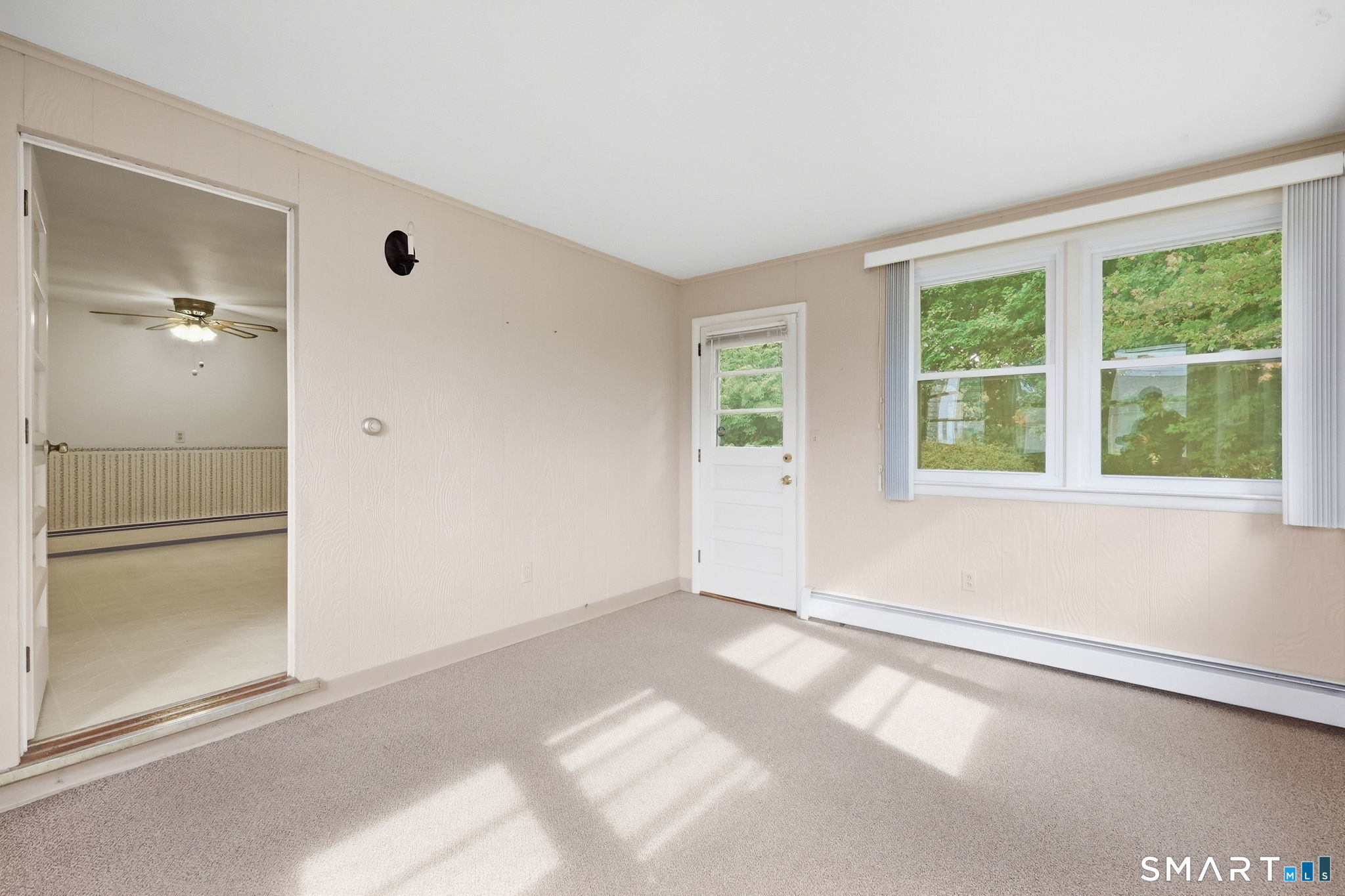 Empty carpeted room with broad windows, a white exterior door, and sunlight across the floor.