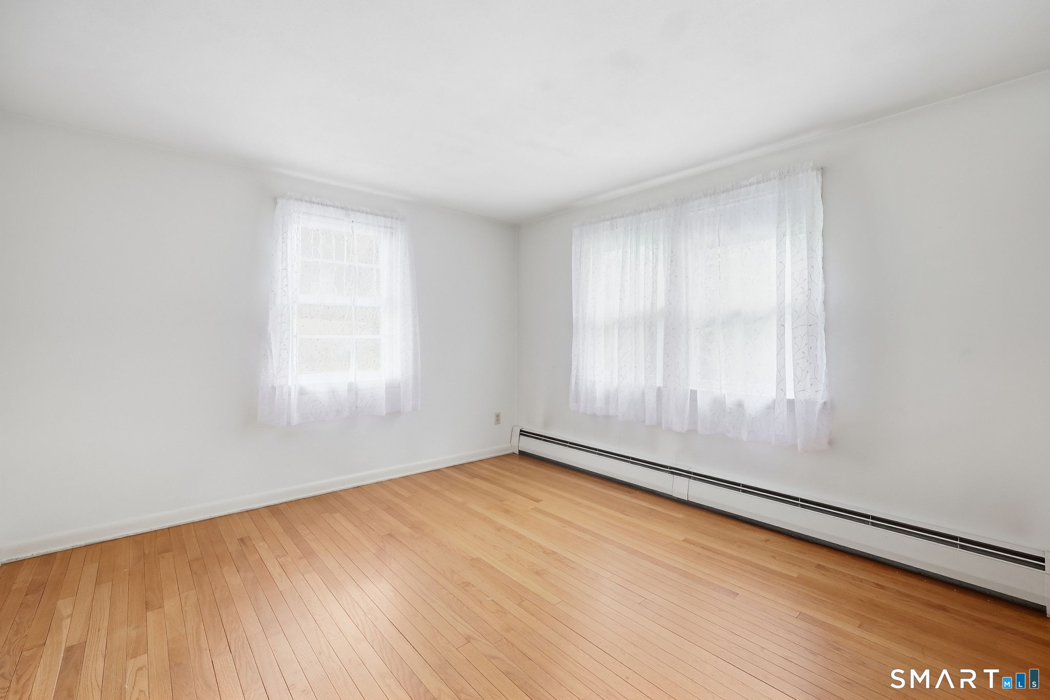 Empty room with hardwood floors, two curtained windows, and a long baseboard heater.