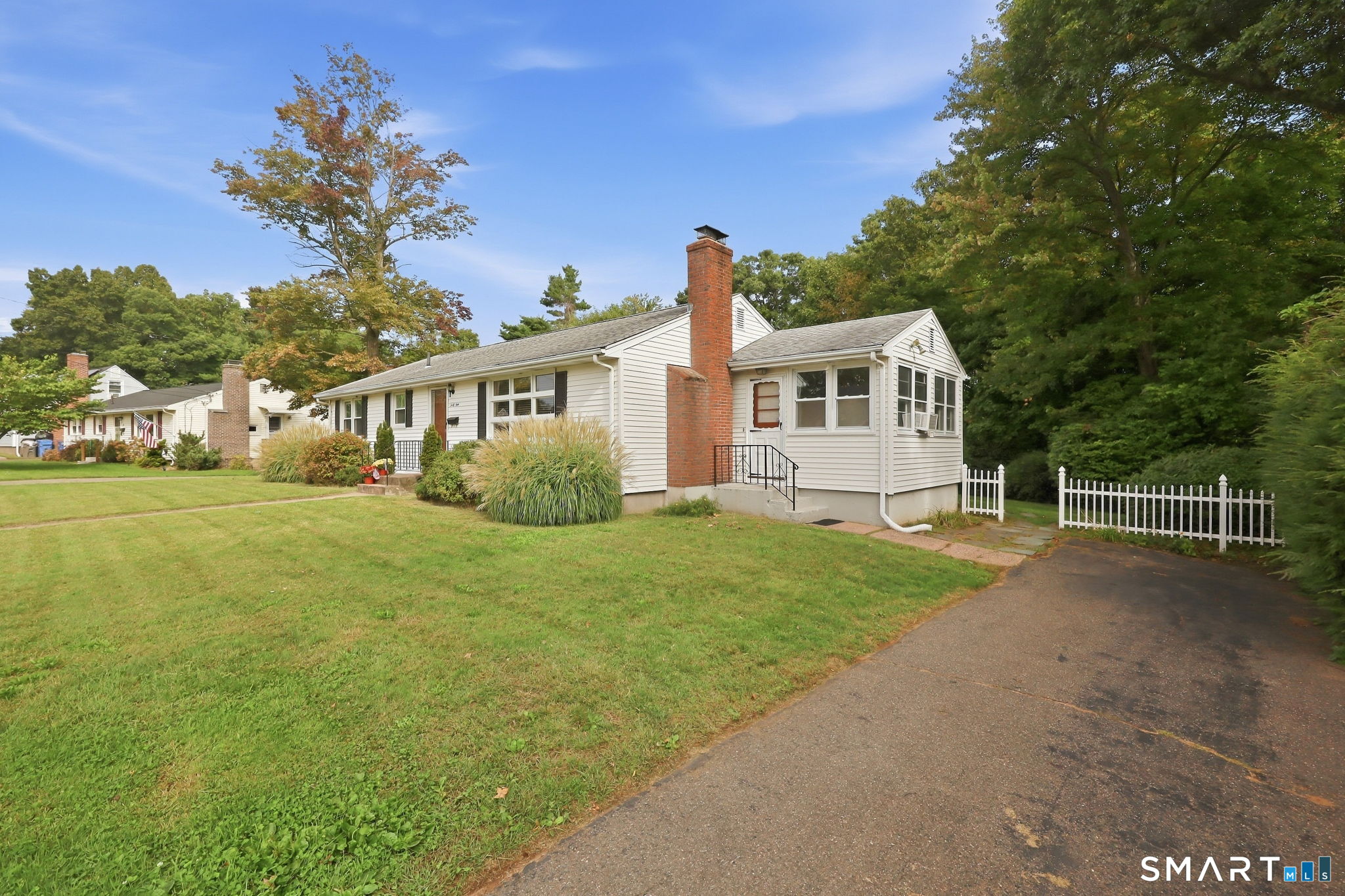 Single-story white house with a brick chimney, enclosed porch, broad lawn, and white picket fencing.
