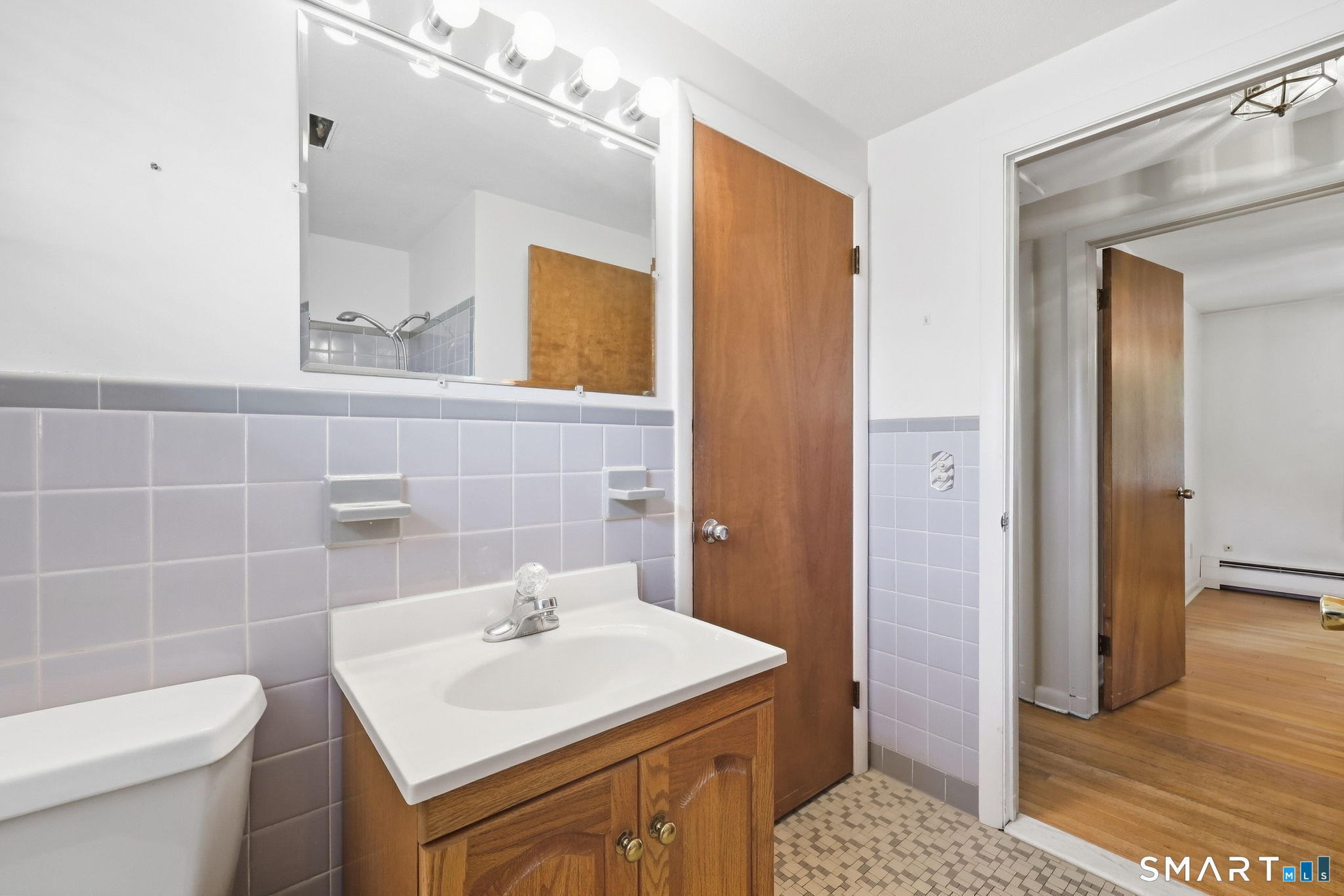 Bathroom with lavender tile, a wood vanity, and a mirror with vanity lights; doorway opens to a hardwood-floored room.