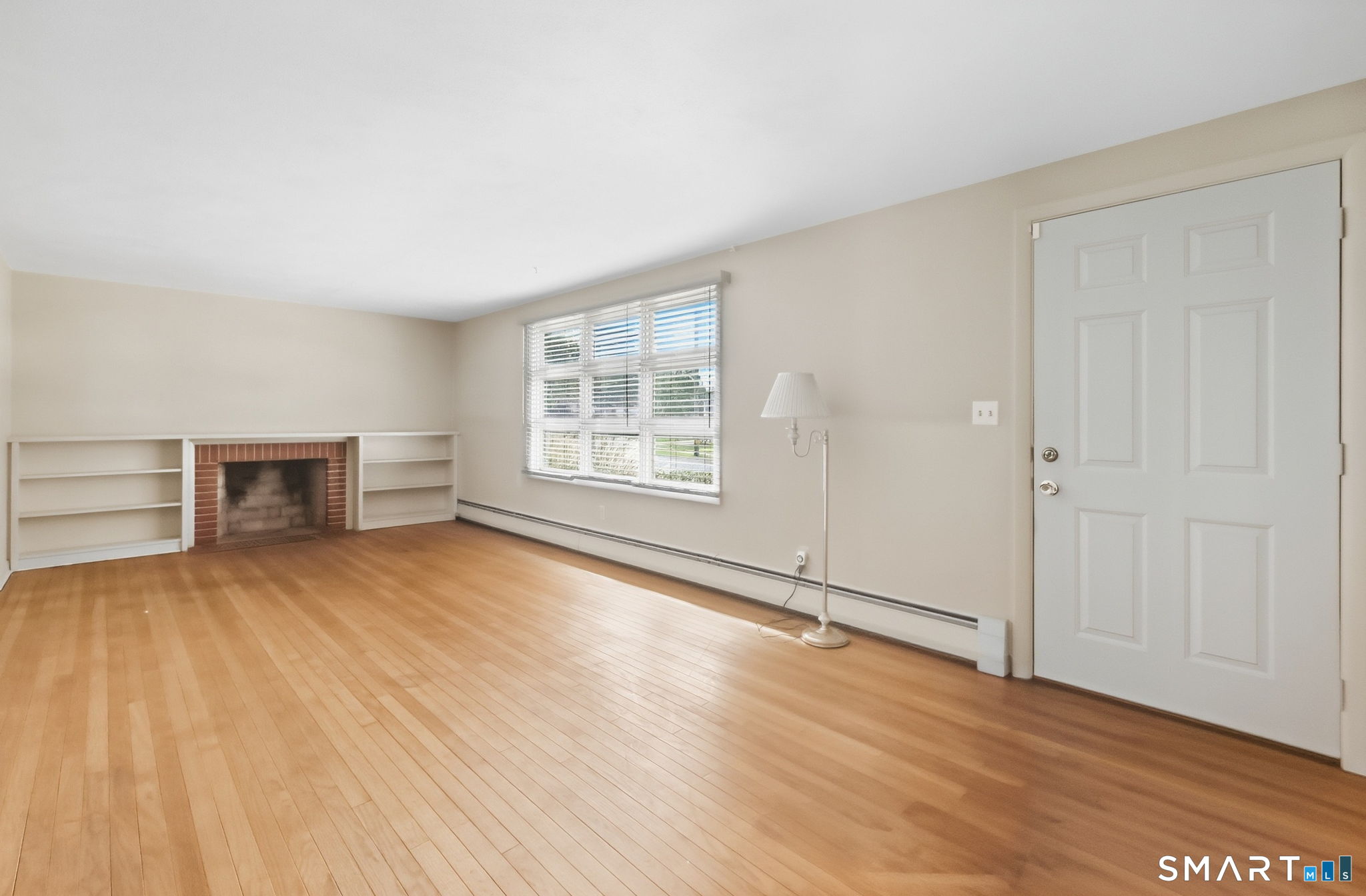 Vacant living room with hardwood floors, brick fireplace, built-in shelving, and a wide window.