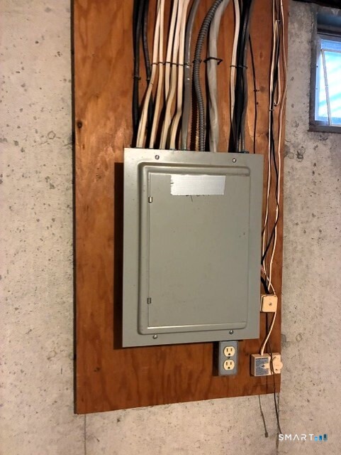 Electrical panel and exposed wiring mounted on plywood against a concrete basement wall.