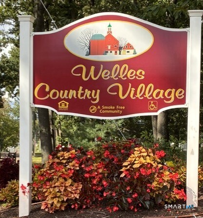 Welles Country Village entrance sign above colorful autumn shrubs.