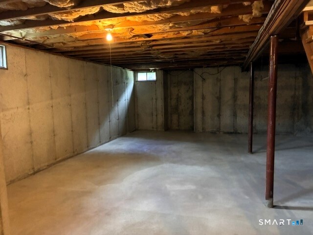 Unfinished basement with concrete floor, exposed ceiling joists, support posts, and small windows.