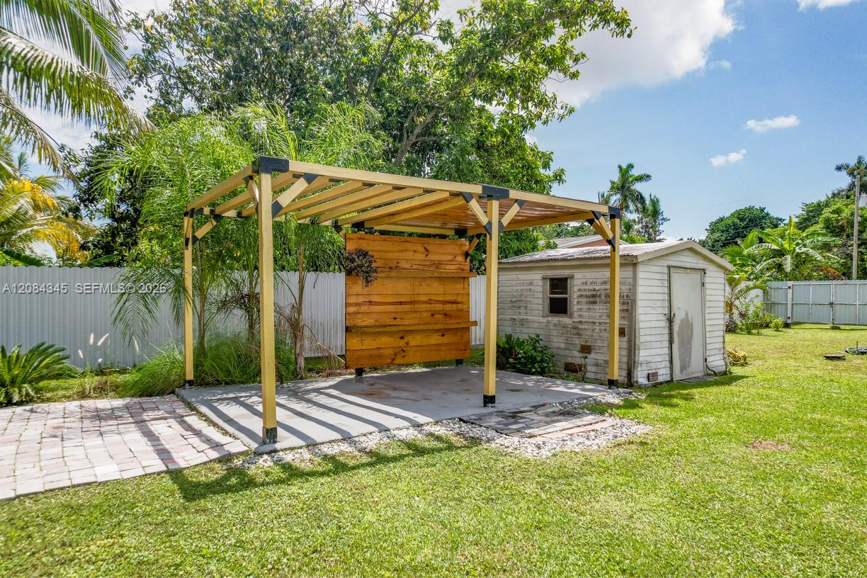 Backyard patio beneath a wooden pergola, beside a small shed and tropical landscaping.
