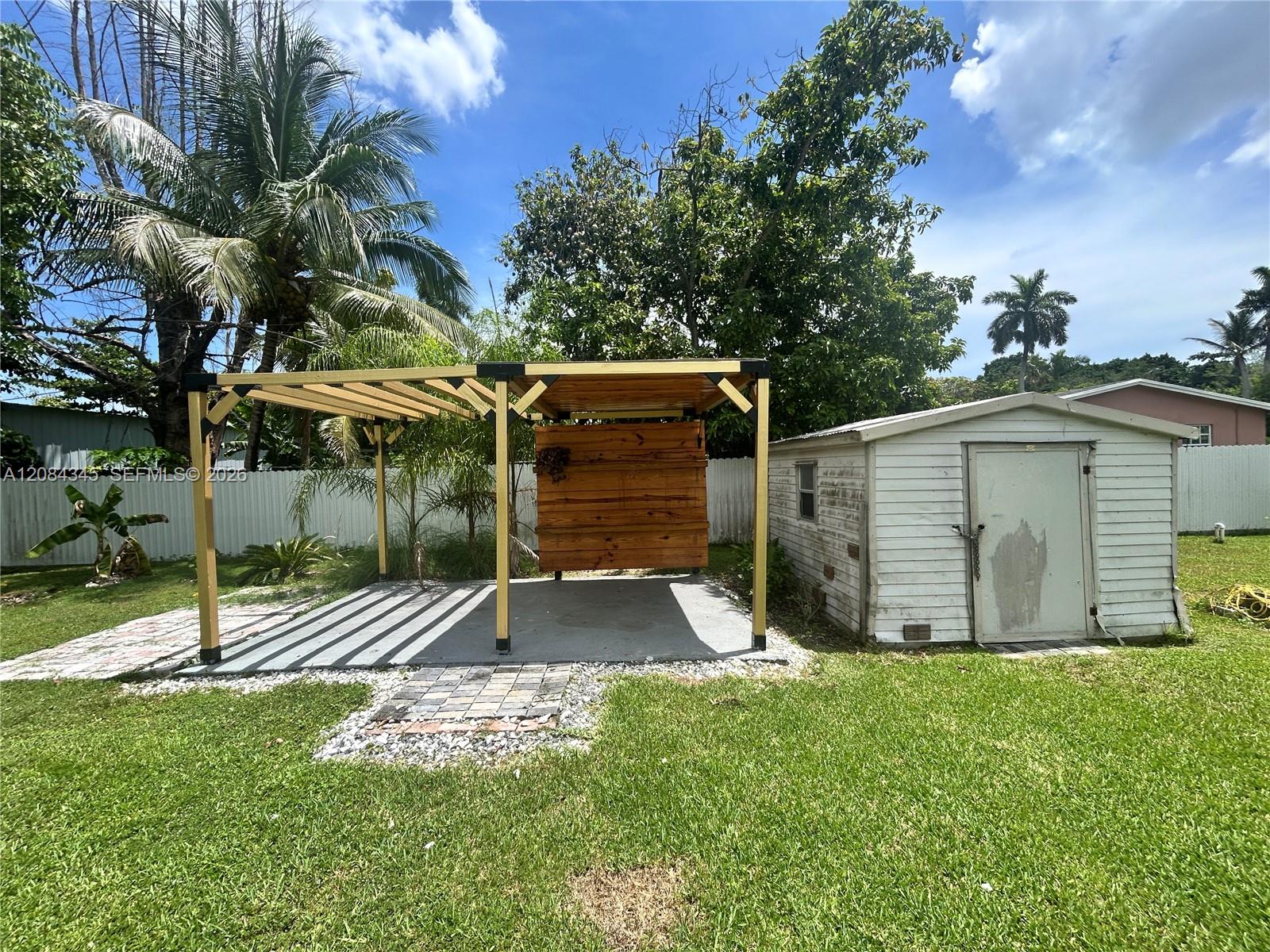 Backyard with a wooden pergola, paved patio, white storage shed, and tropical trees.