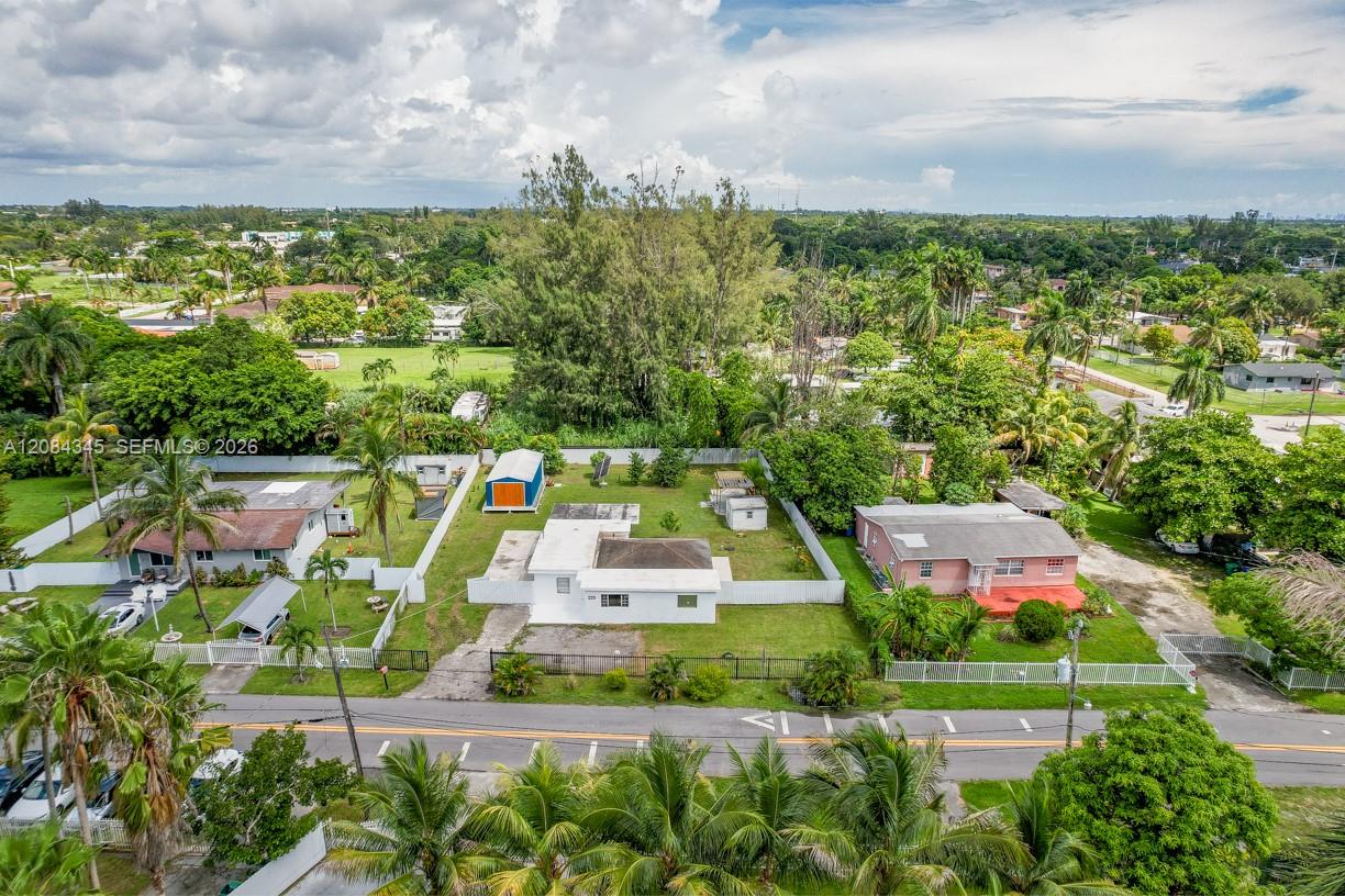 Aerial view of a white single-story house with a fenced grassy yard and outbuildings among palm trees.