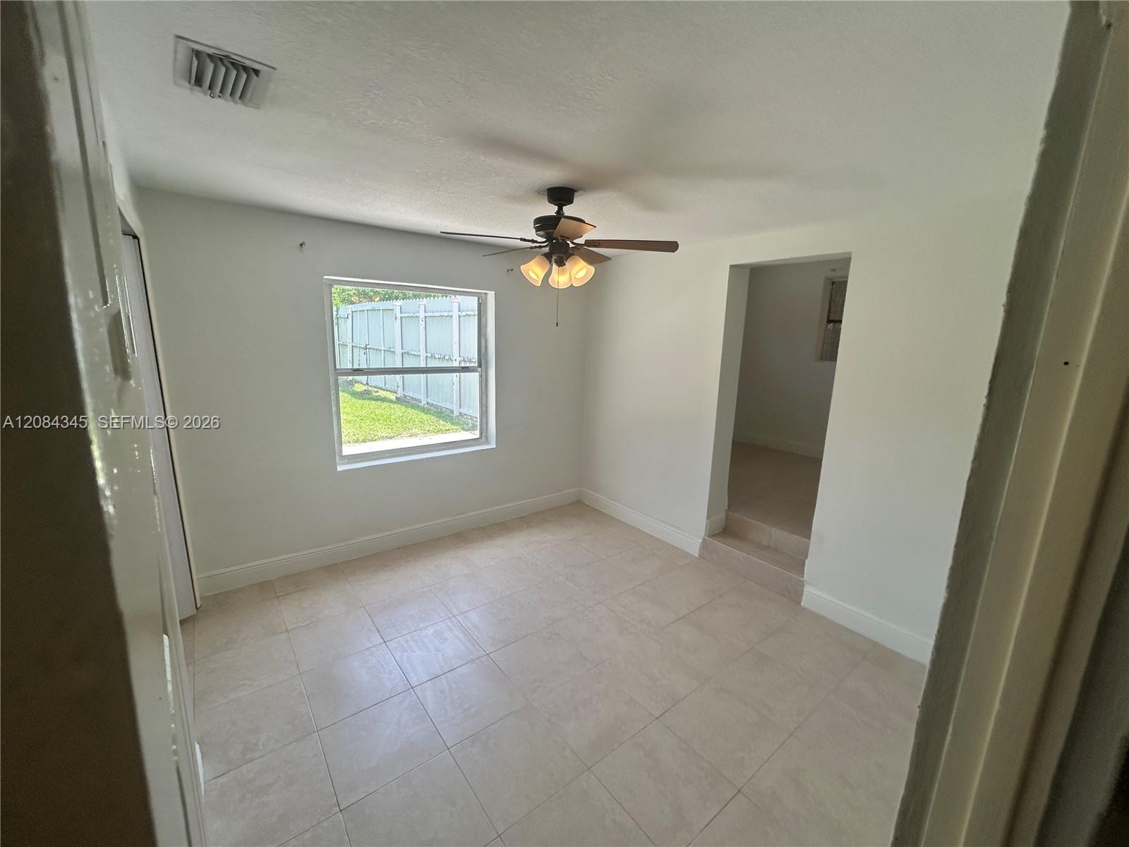 Empty tiled room with a ceiling fan and a window overlooking a fenced yard.