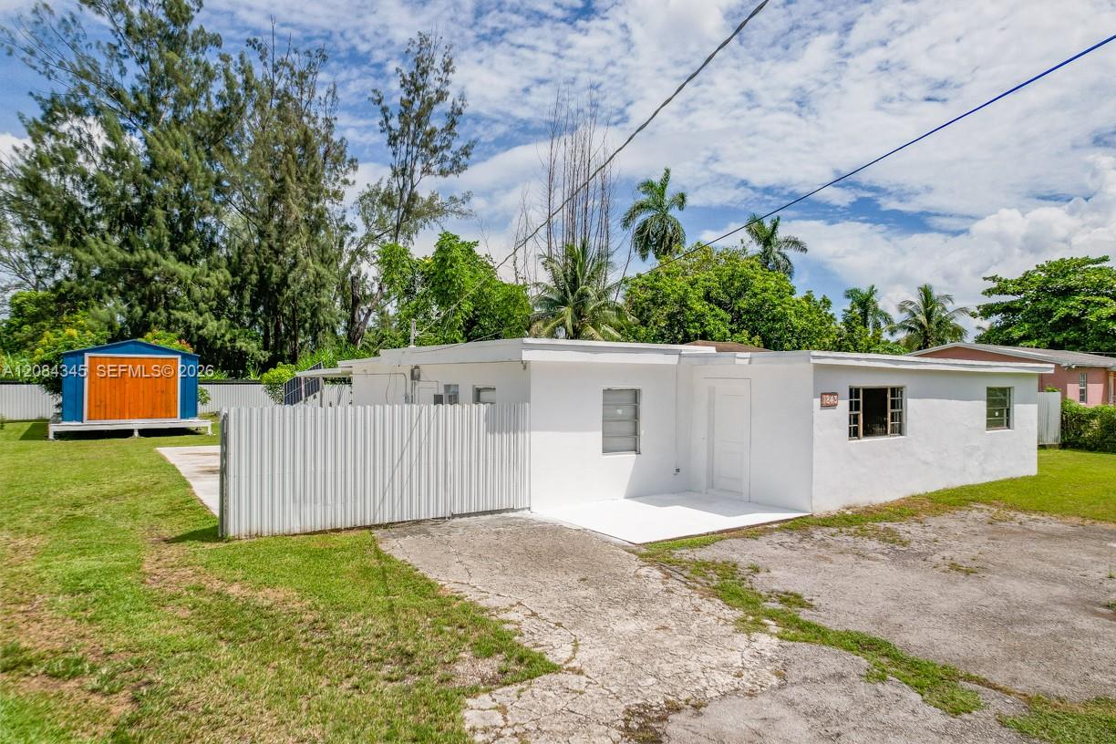 White single-story home with a flat roof, fenced yard, colorful shed and cracked driveway.