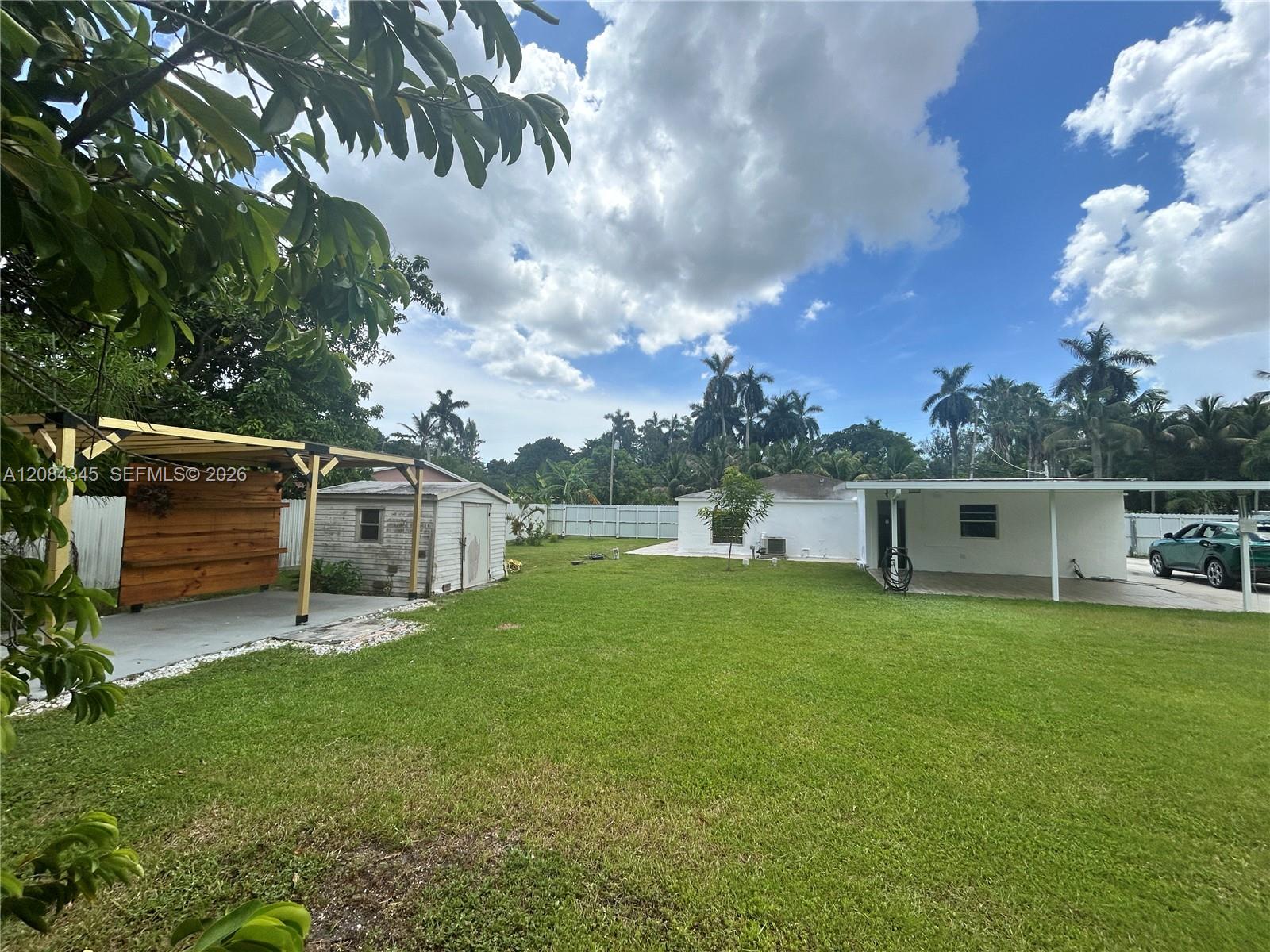 Large grassy backyard with a wooden covered patio, shed, single-story home, and carport.