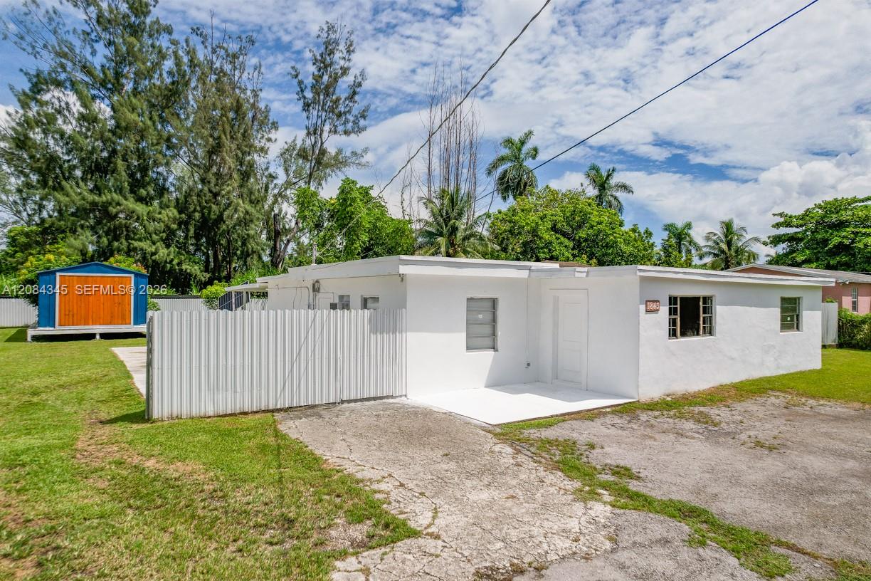 White single-story home with a fenced lawn, colorful shed, palm trees, and cracked driveway.