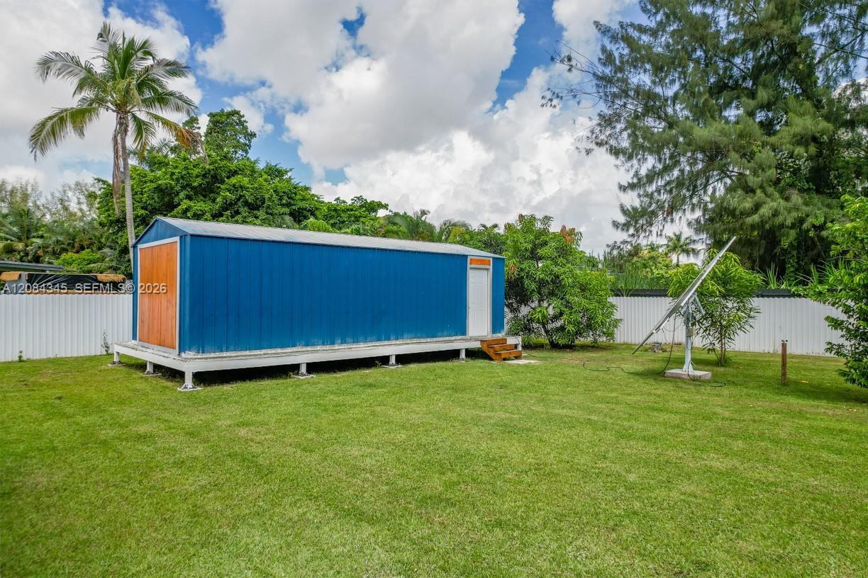 Blue detached outbuilding raised above a grassy yard behind a white fence.