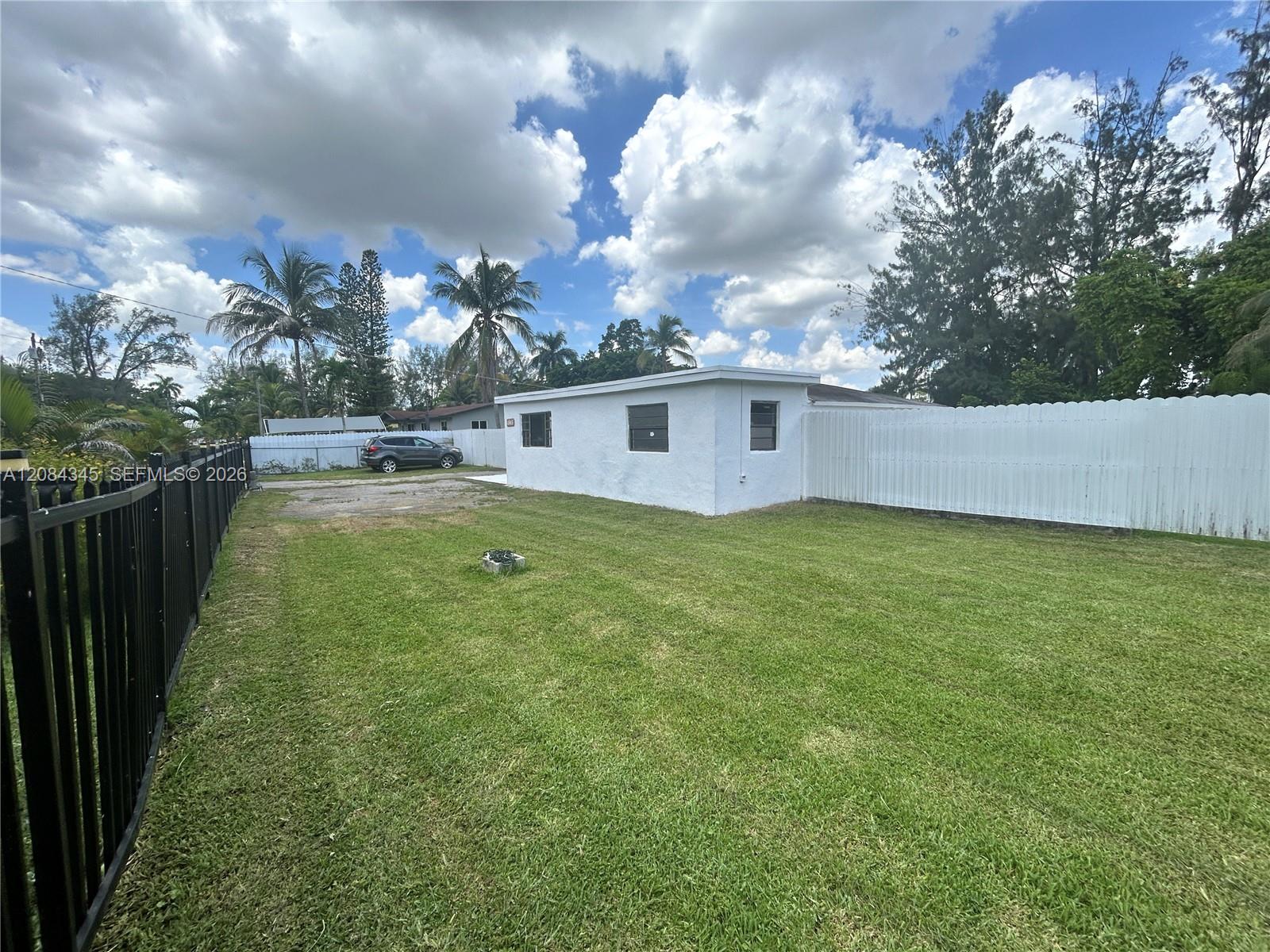 Grassy yard beside a white single-story house, enclosed by white and black fences with palm trees beyond.