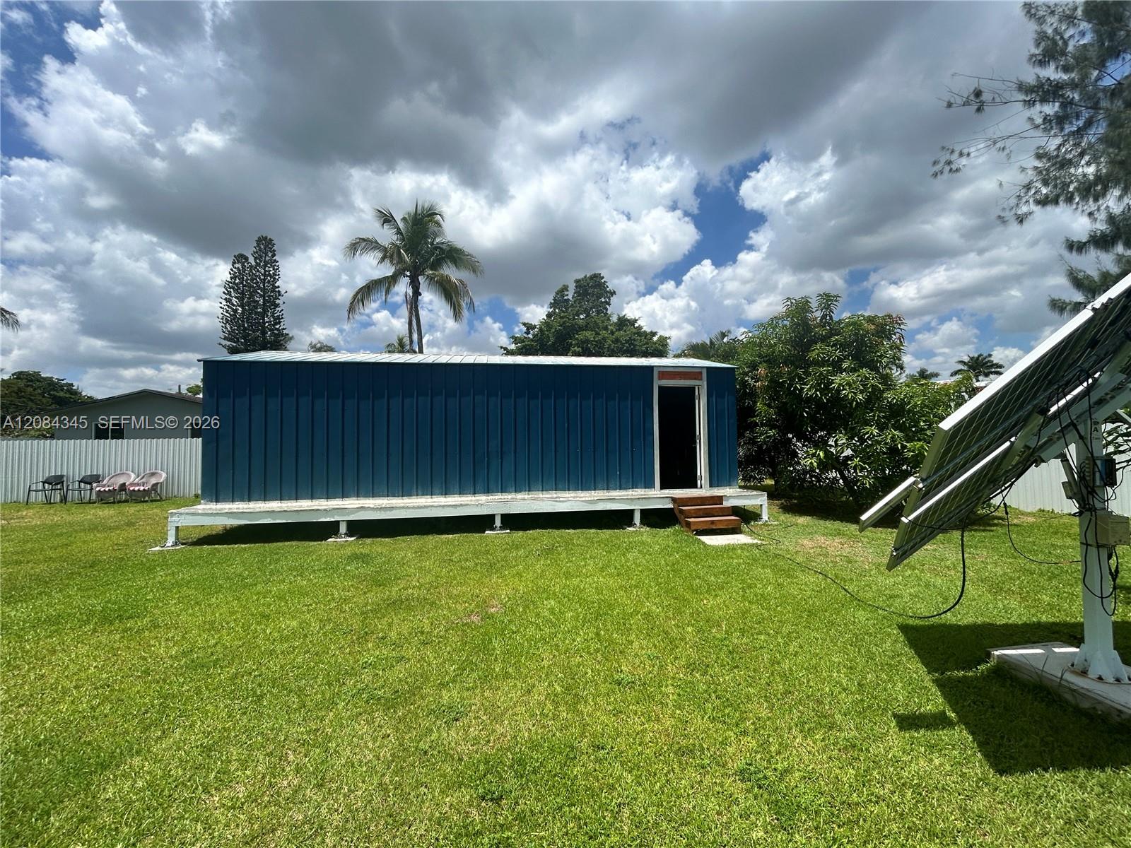 Blue raised structure with an open doorway on a grassy lot beside solar panels.