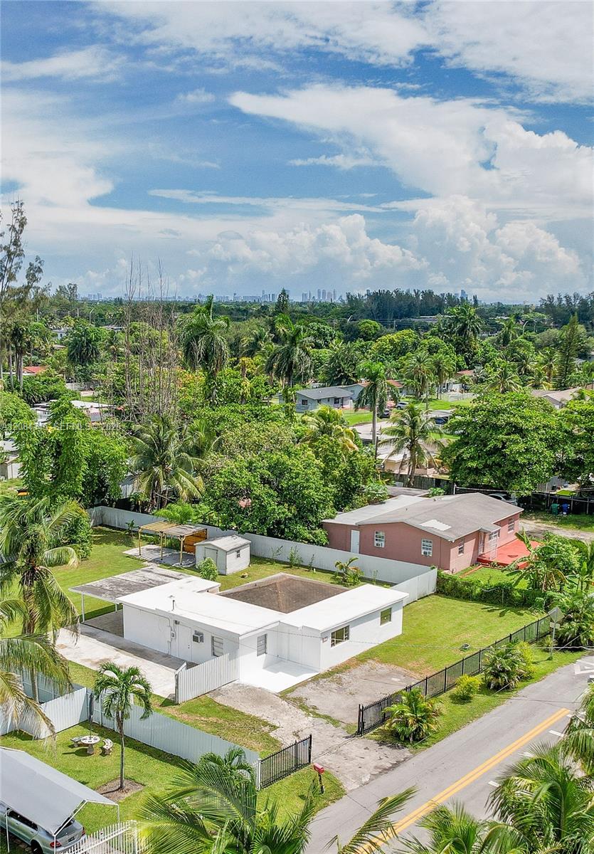 Aerial view of a white single-story house with a fenced yard, palm trees, and the Miami skyline in the distance.