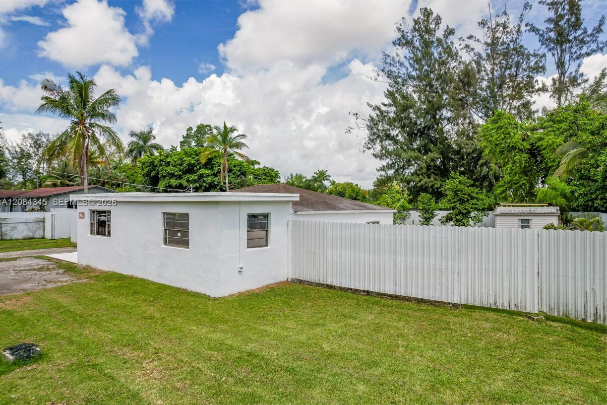 Single-story white house beside a tall privacy fence, with a broad lawn and palm trees.