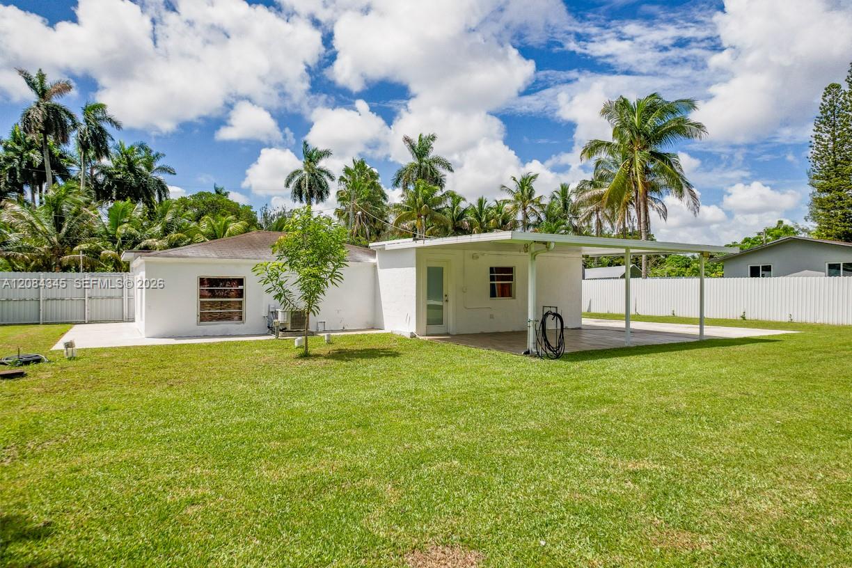 Rear of a single-story white house with a covered carport, broad lawn, and palm trees.