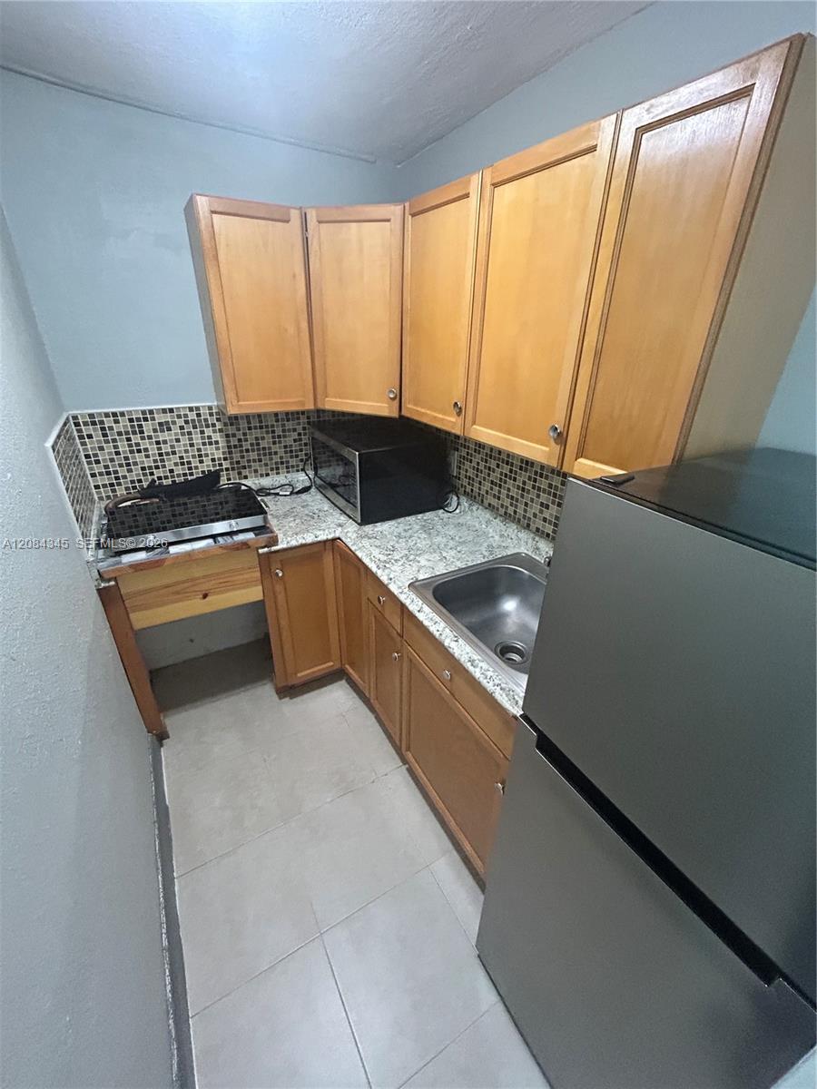 Compact kitchen with light wood cabinets, mosaic backsplash, and a stainless-steel refrigerator beside the sink.