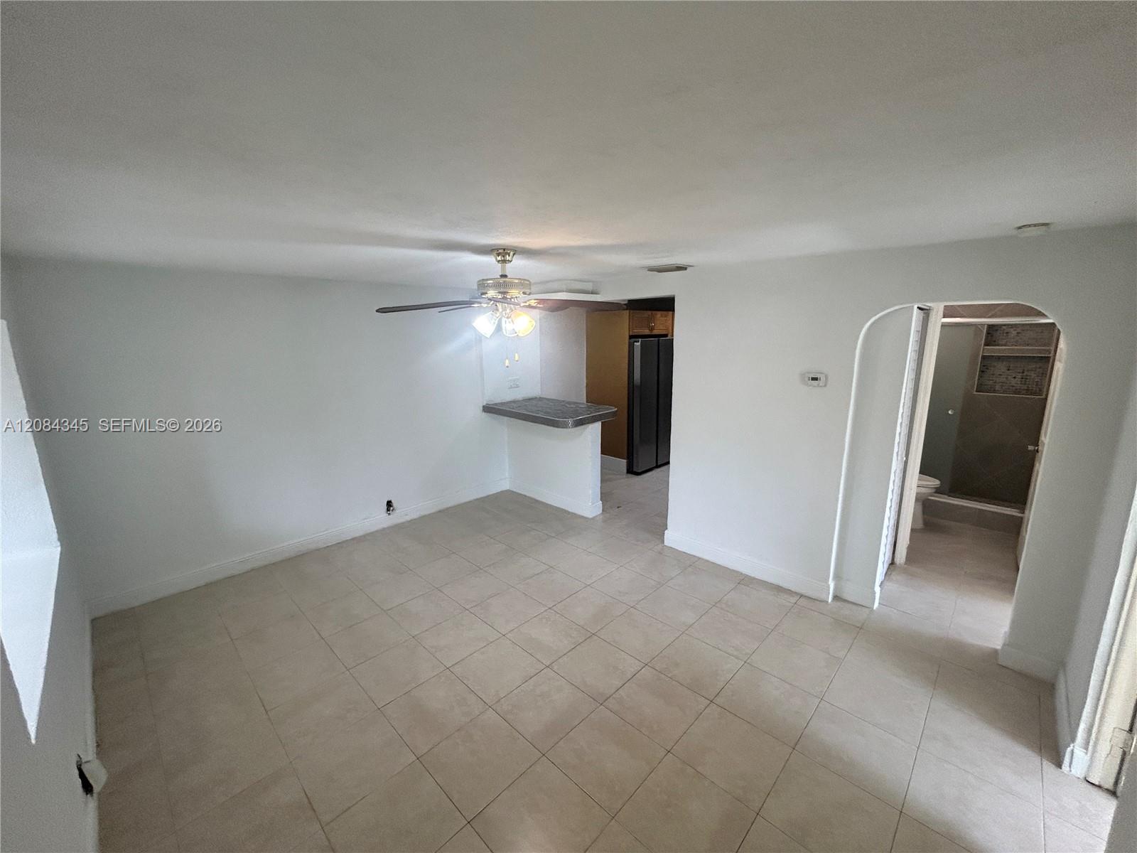 Unfurnished living area with tiled floors, a ceiling fan, kitchen pass-through, and arched bathroom doorway.