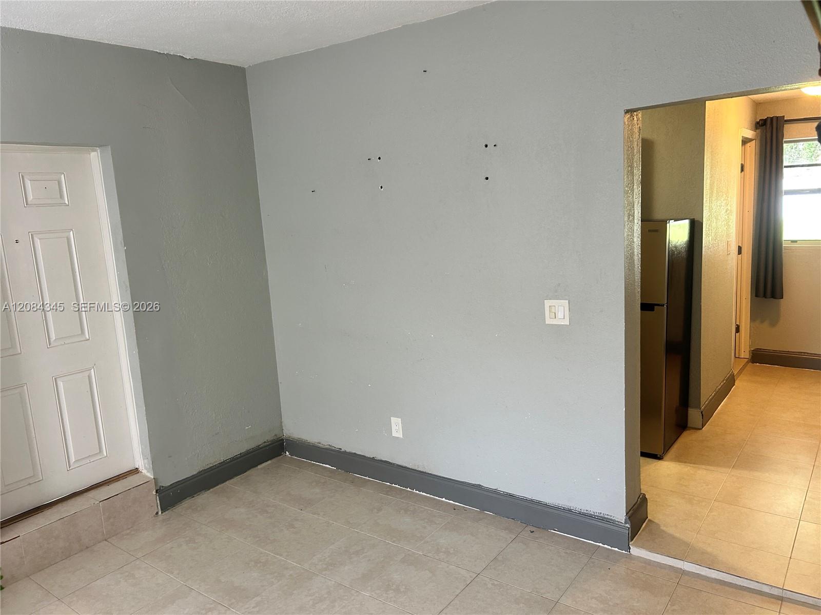 Unfurnished tiled room with gray walls, a white door, and an opening to a kitchen area.