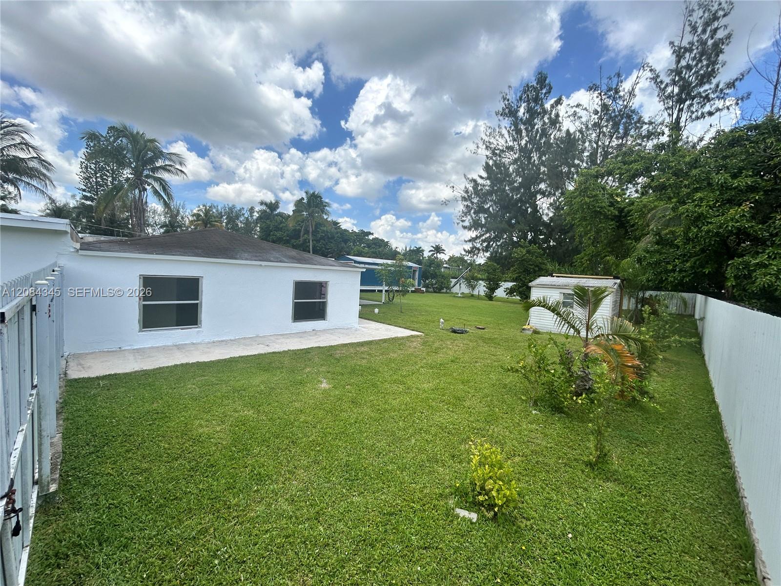 Fenced grassy backyard behind a white house, with a paved patio and small shed.