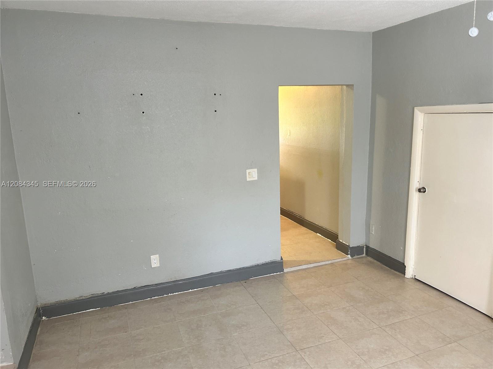 Empty gray-walled room with beige tile flooring, a doorway, and a closed white door.
