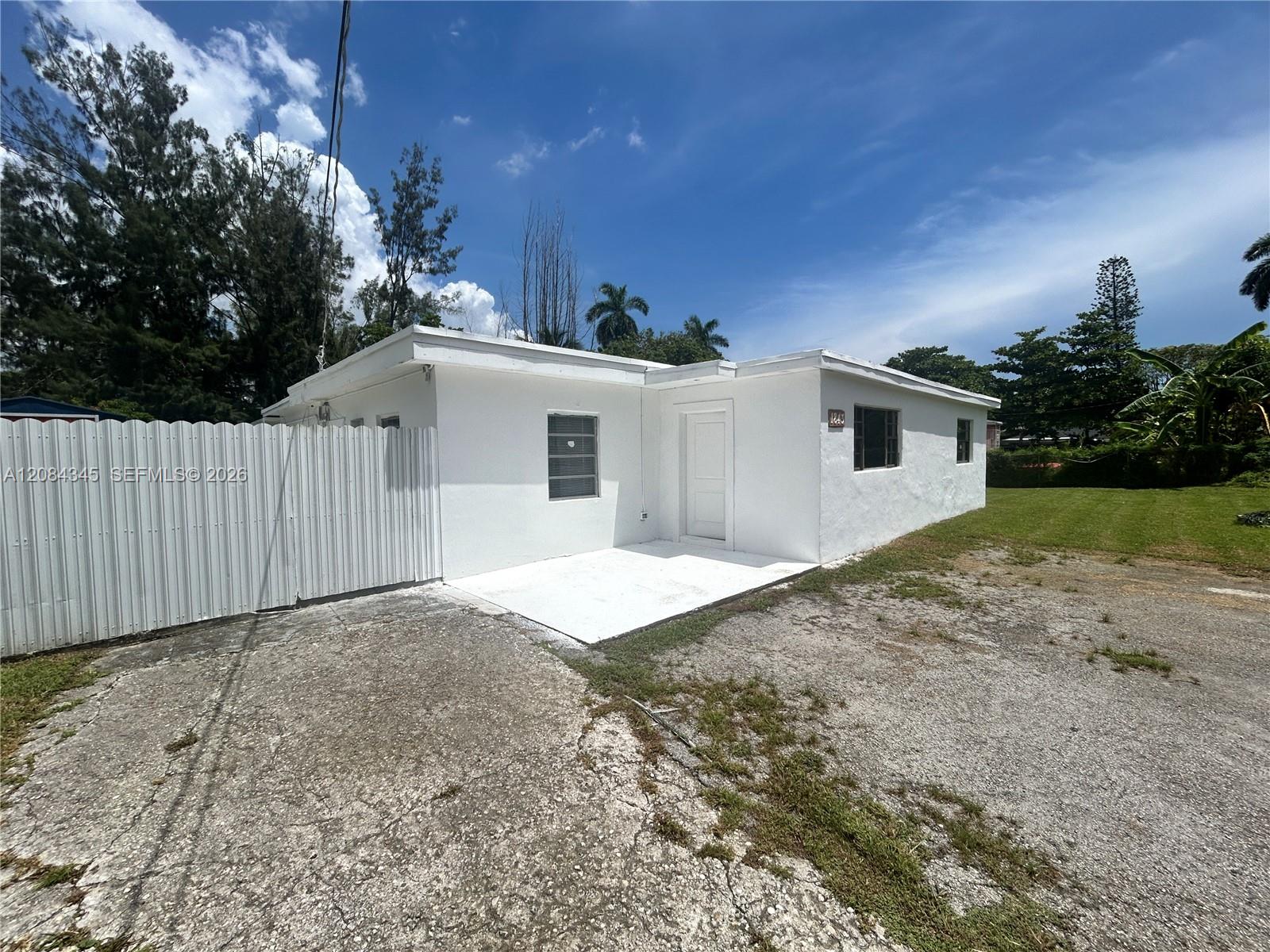 White single-story home with a flat roof, corrugated side fence, paved driveway, and grassy yard.