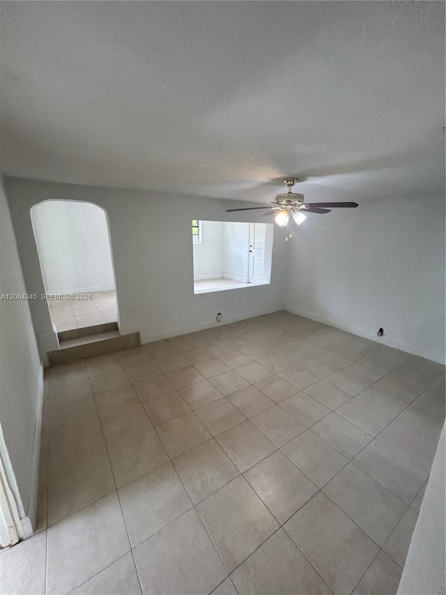 Empty tiled living room with a ceiling fan and arched doorway to an adjoining room.