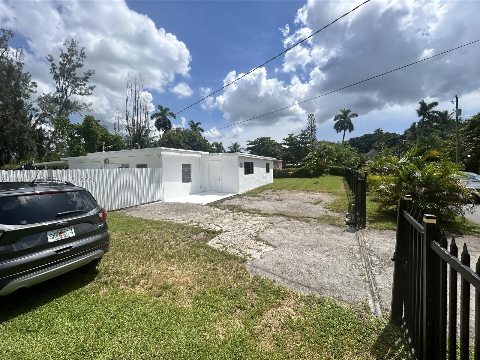 Single-story white house beside a cracked driveway, grassy yard, and black metal fence.