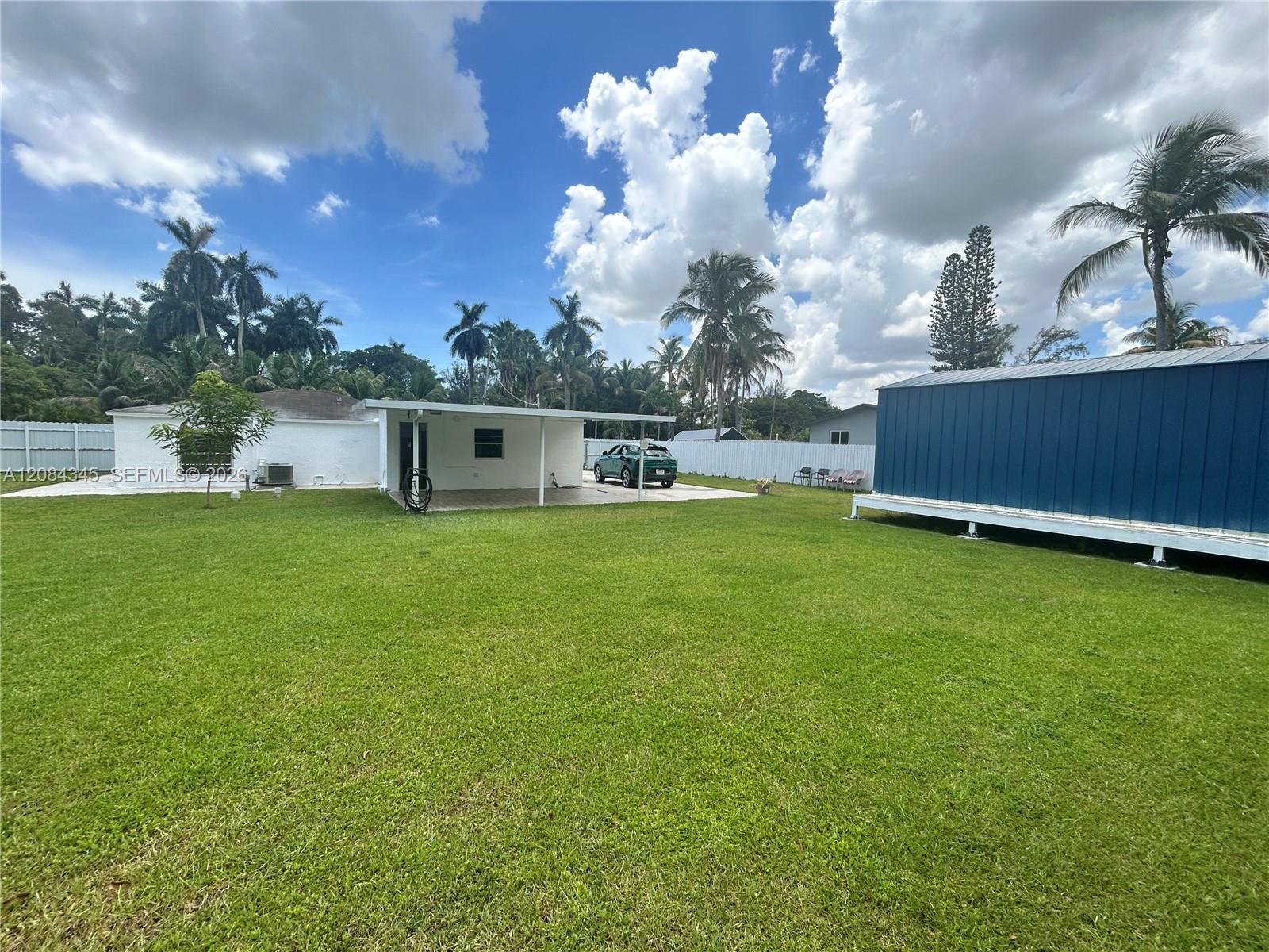 Rear yard with broad lawn, covered carport, parked vehicle, and large blue outbuilding.