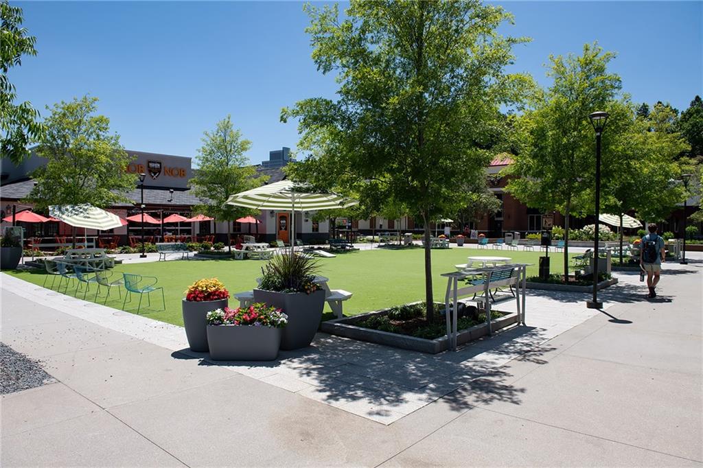 Outdoor plaza courtyard with artificial turf, shaded dining tables, large planters, and surrounding storefronts.