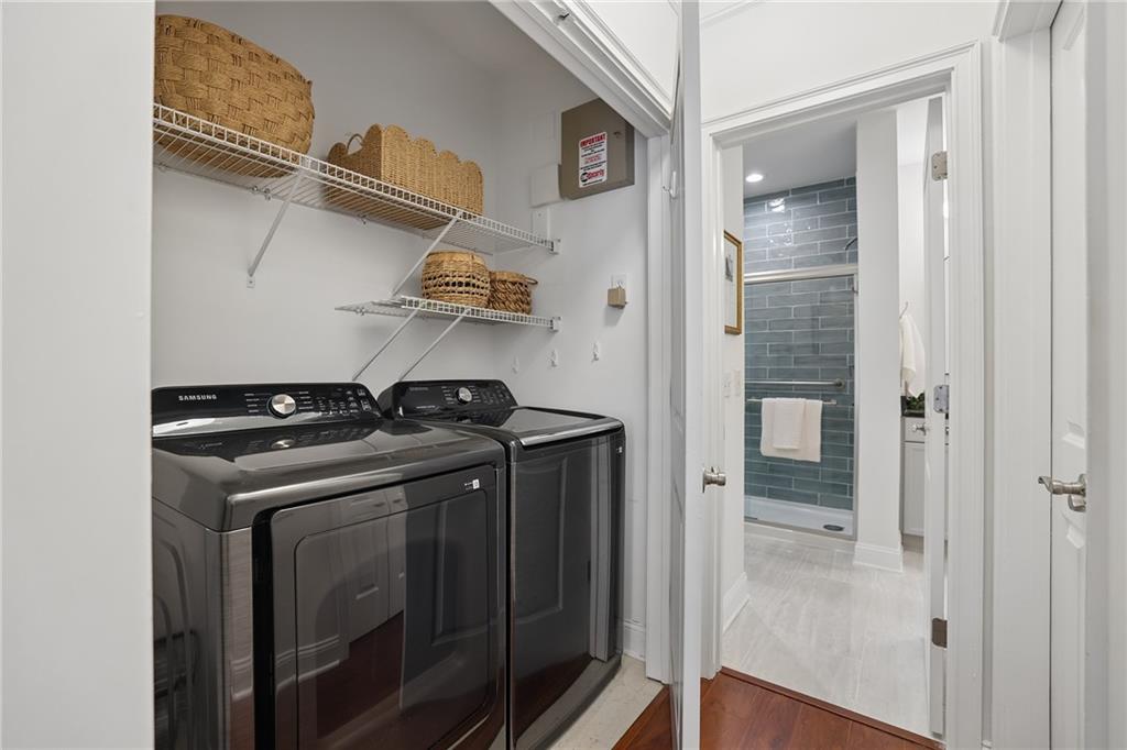 Laundry room with dark Samsung washer and dryer, woven baskets, and a tiled shower visible through the doorway.