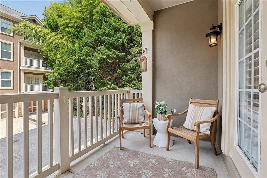 Covered balcony with cushioned chairs, a small table, and a view of trees and apartments.