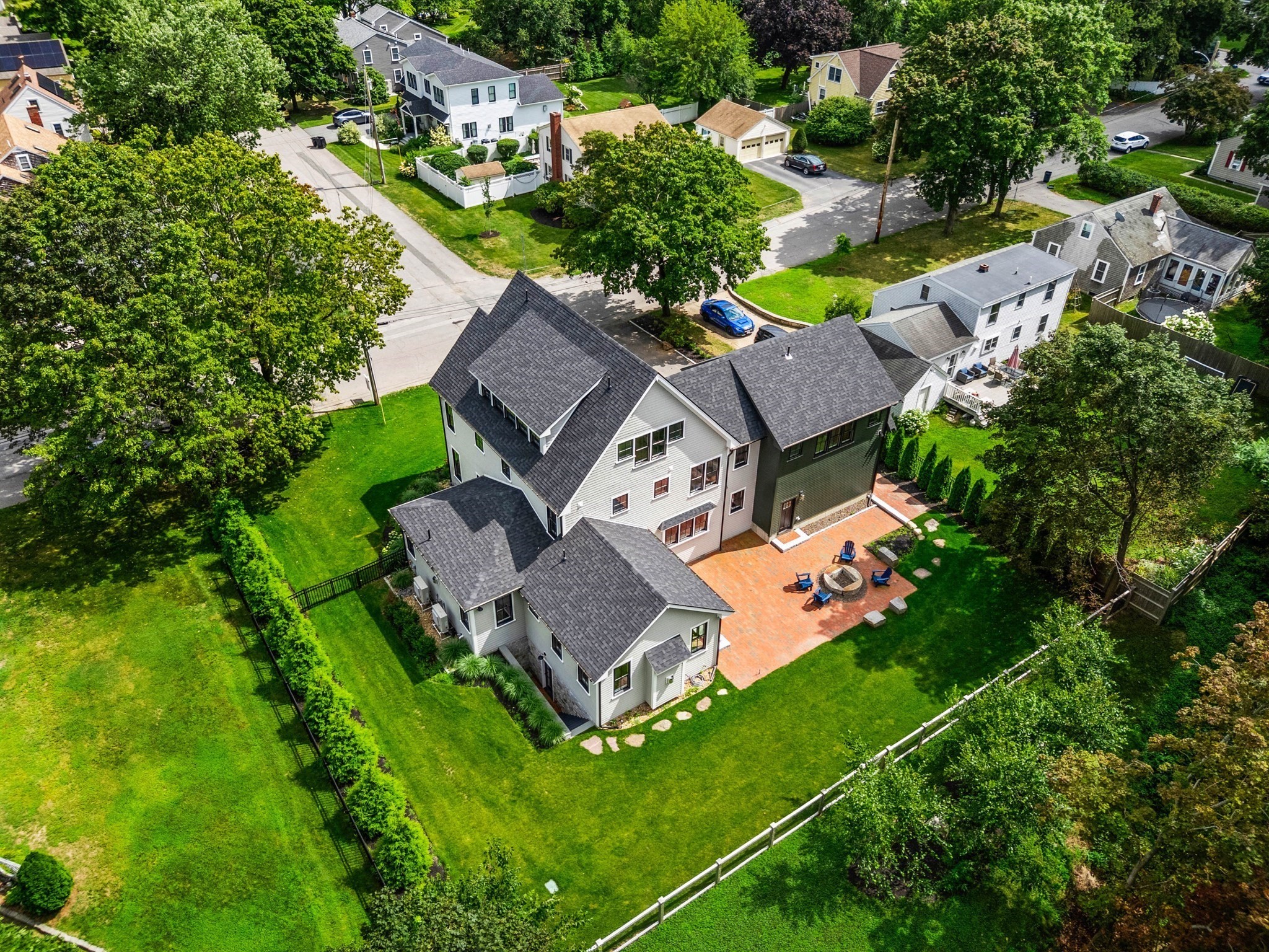 Aerial view of a multi-section house with a brick patio, circular fire pit, and expansive fenced lawn.