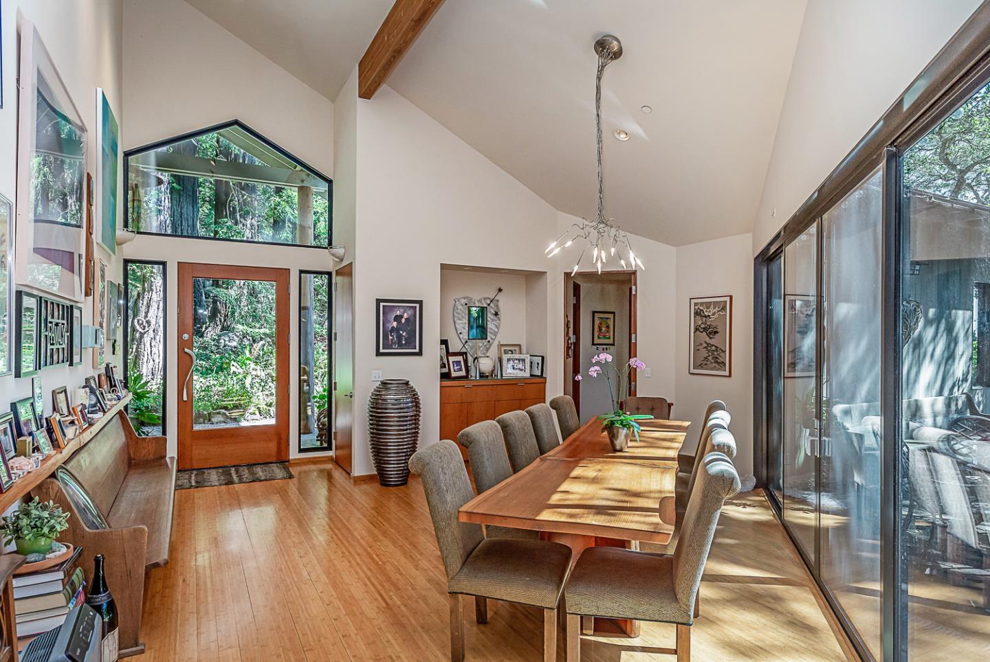Dining room with a long wooden table, vaulted ceiling, and tall windows overlooking trees.