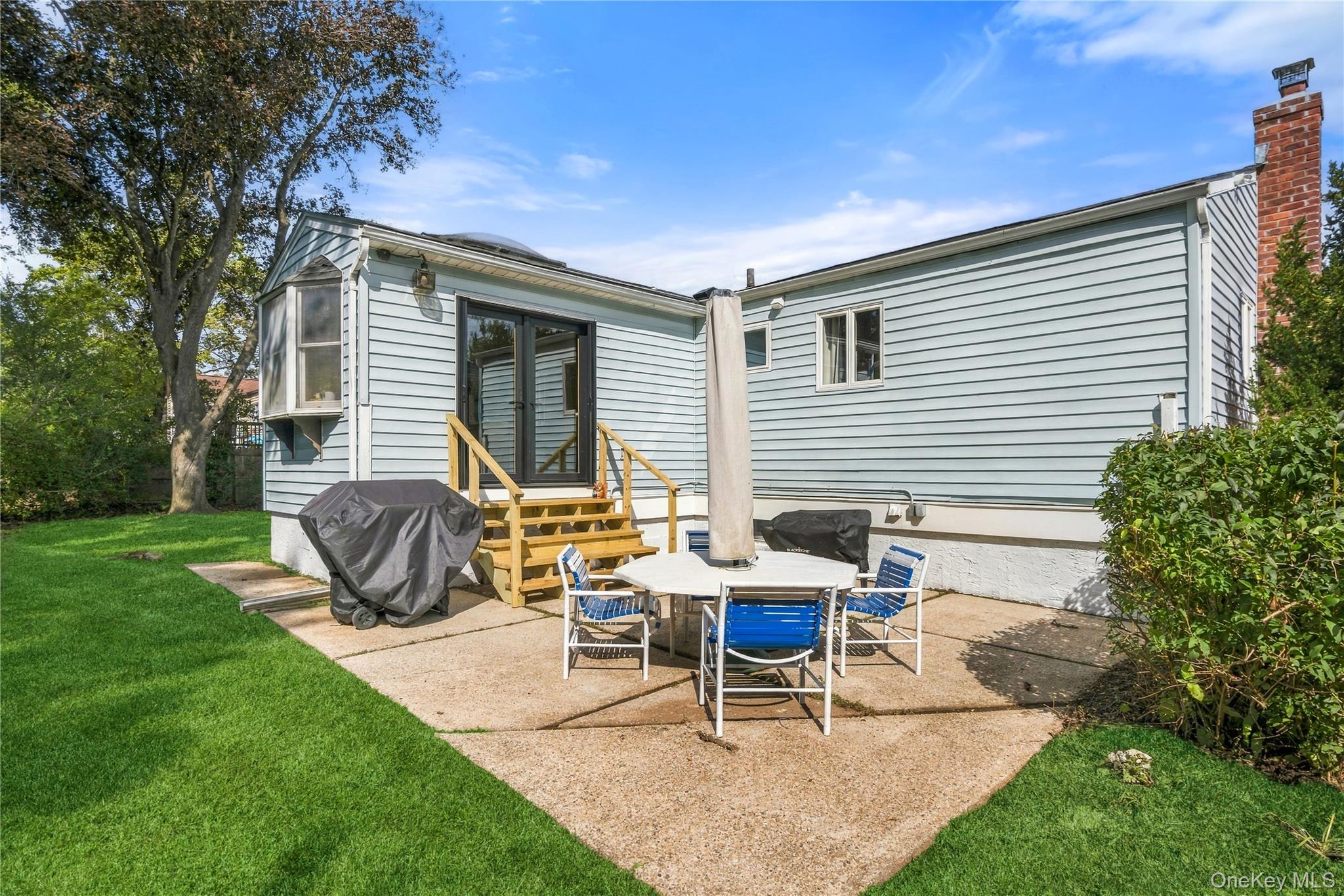 Backyard patio with blue dining chairs, wooden steps, and a green lawn beside the house.