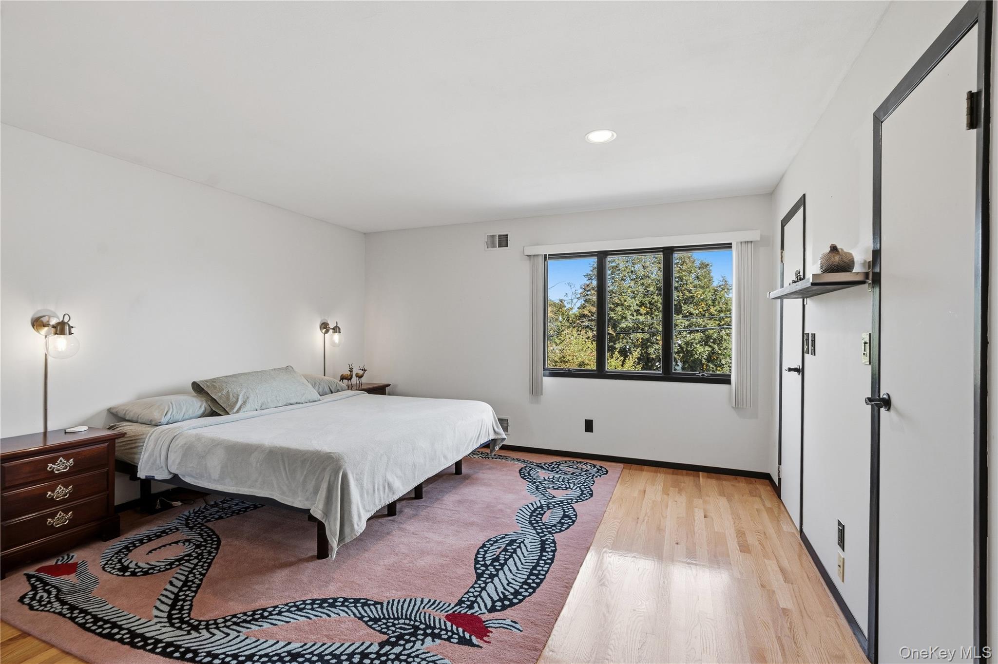 Furnished bedroom with a large bed, patterned rug, hardwood floors, and a tree-facing window.