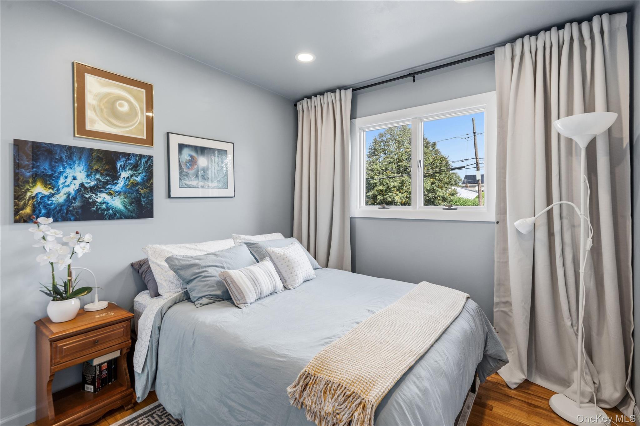 Bedroom with pale blue bedding, framed artwork, and tall curtains beside a sunny window.
