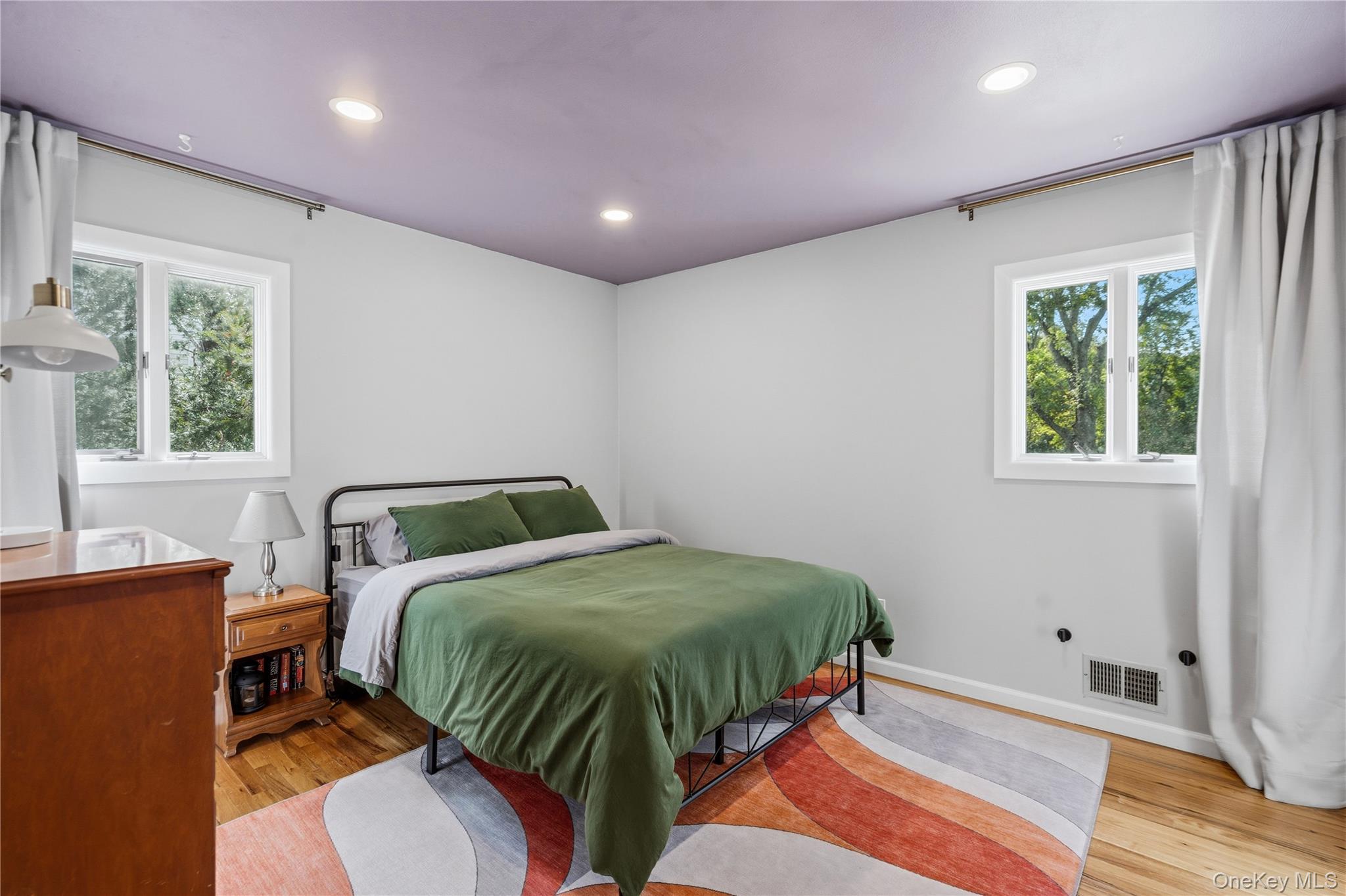 Bedroom with green bedding, two curtained windows, hardwood floors, and a colorful geometric rug.
