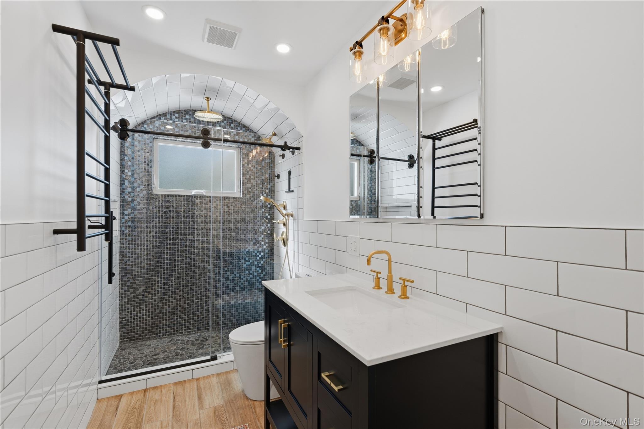 Bathroom with an arched blue-gray mosaic shower, black vanity, and gold fixtures.