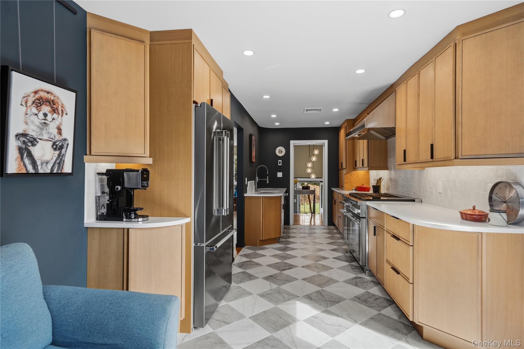 Long kitchen with light wood cabinets, stainless steel appliances, and checkerboard tile flooring.