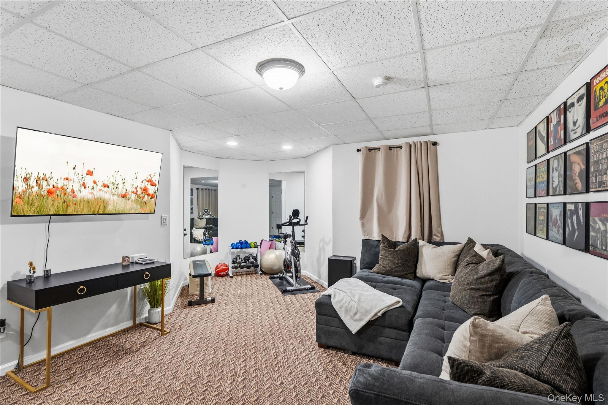 Recreation room with a gray sectional, wall-mounted television, exercise bike, weights, and framed music posters.
