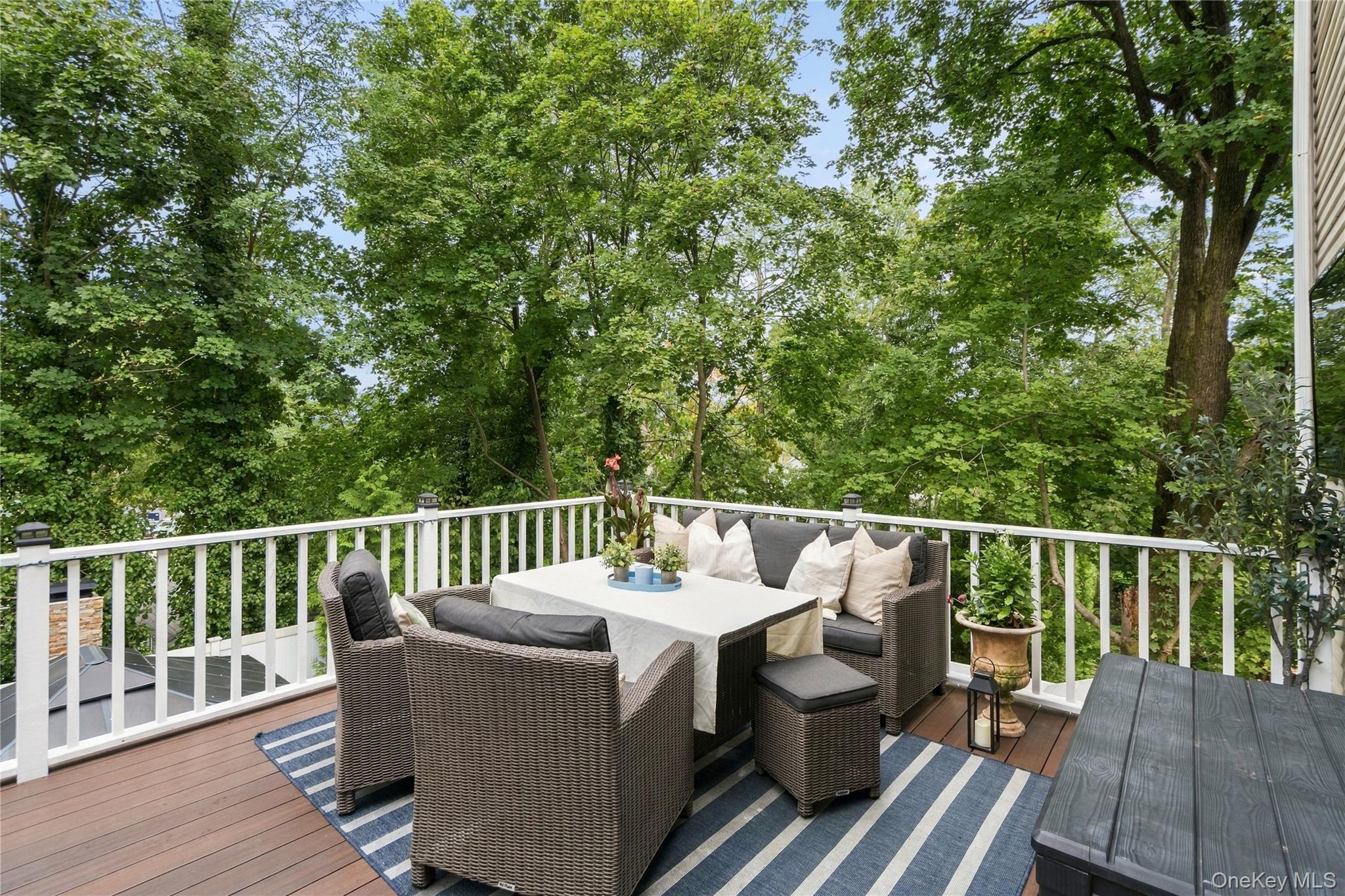 Furnished deck with wicker dining set and striped rug, surrounded by leafy trees.