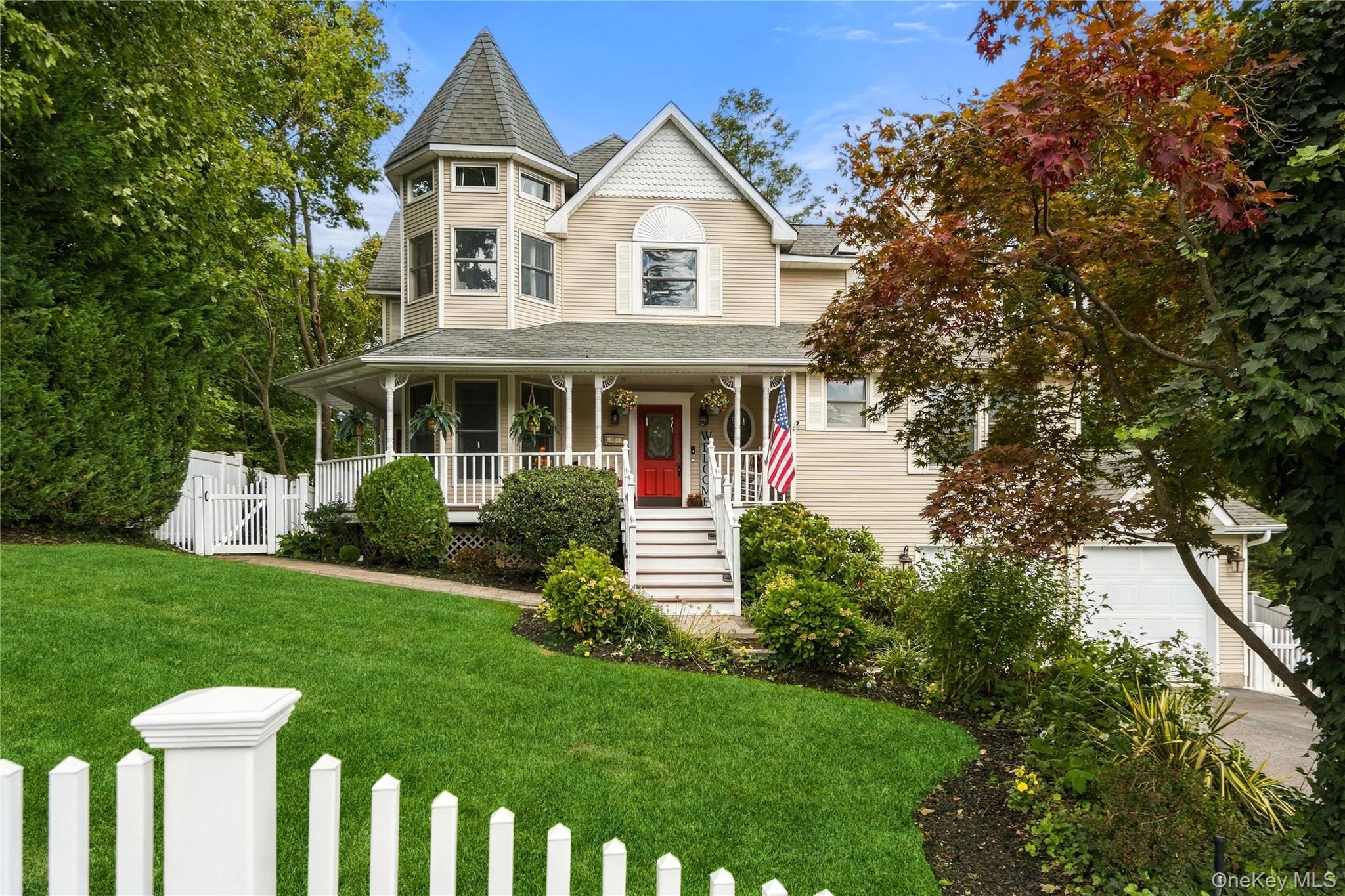 Beige home with a turret, red front door, and covered porch behind a white picket fence.